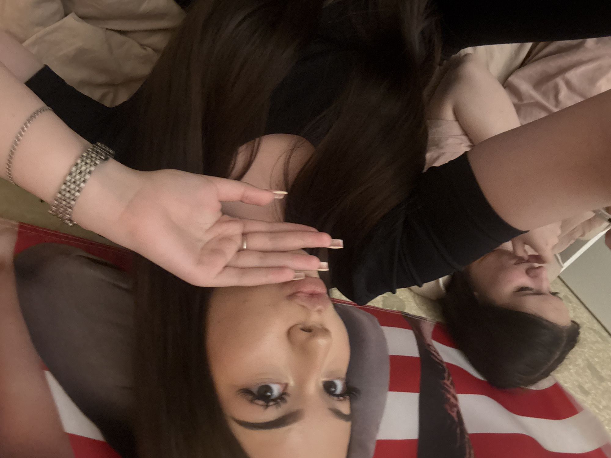 This media file features two females in a casual indoor setting, likely a bedroom, with the image rotated 90 degrees counter-clockwise.
The primary subject, in the foreground, is a young woman with long, dark hair, lying down and looking directly into the camera. Her head is at the bottom of the frame, resting on a gray pillow, and her body extends upwards. She wears a dark top or dress. Her left wrist is adorned with two metallic bracelets: one broad, composed of multiple rows of square links, and another thinner chain bracelet. Her right hand is raised, with a ring visible on her ring finger and long, light-colored nails covering part of her lower face, suggesting a posed or playful expression. She has prominent eye makeup, including long eyelashes and defined eyebrows, and her lips are slightly parted. Beneath her and extending across a significant portion of the frame is a large fabric item patterned with bold red and white stripes, strongly resembling the American flag.
A second female is partially visible in the background, to the upper right. She also has dark hair and is wearing a light-colored top, lying down and looking away from the camera. The background consists of light-colored, possibly beige or tan, bedding. The overall activity depicts a relaxed moment, possibly a selfie or casual photo session. There is no visible text in the image. The location context for this media file is Naberezhnye Chelny, Russia.
