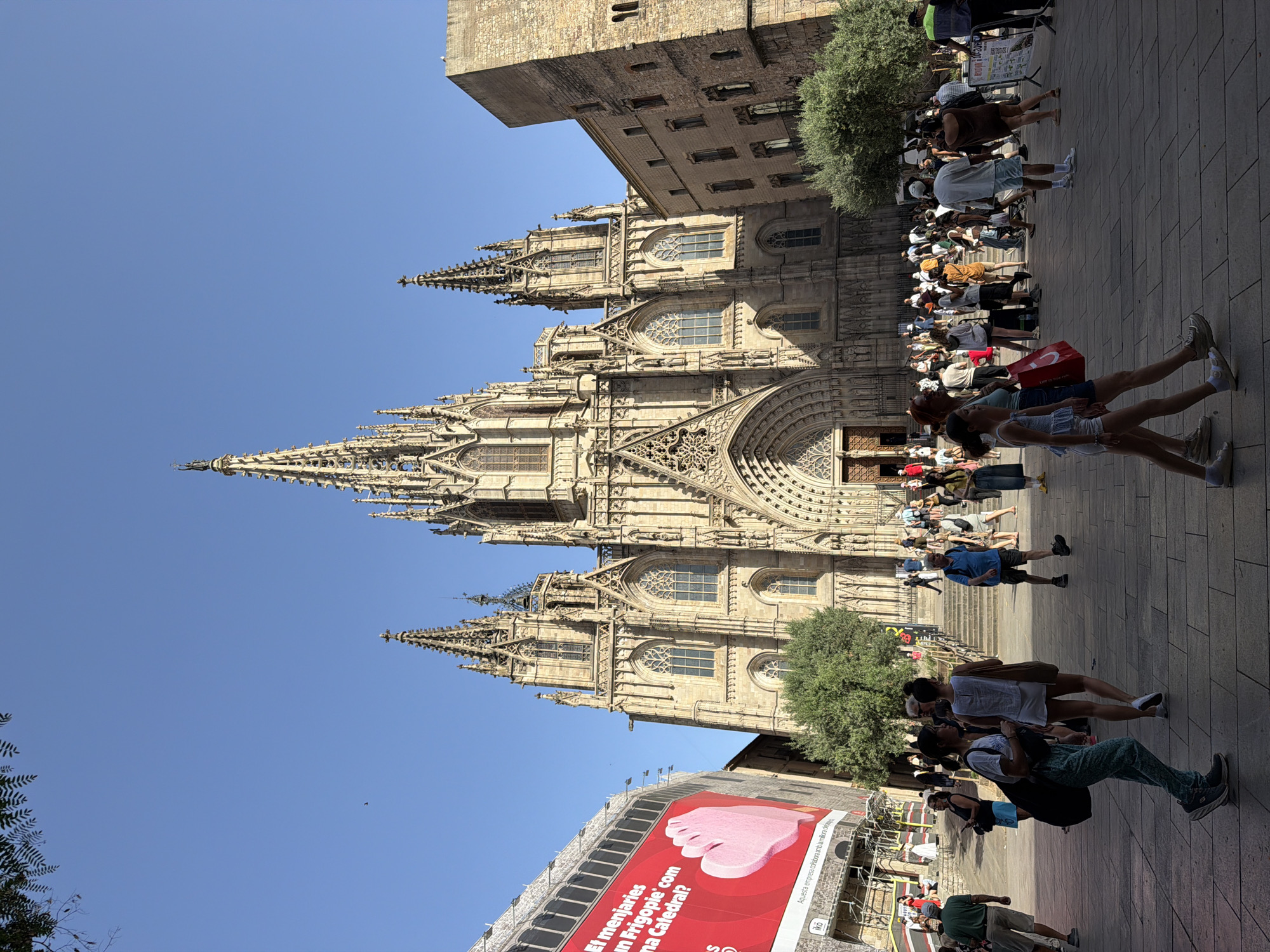 This is an eye-level, medium shot of the Barcelona Cathedral, also known as the Cathedral of the Holy Cross and Saint Eulalia, in Barcelona, Spain. The cathedral is a Gothic structure with intricate spires and arched doorways. The facade is made of light-colored stone. In front of the cathedral is a wide, paved plaza filled with numerous people walking around and congregating. Some individuals are dressed casually, suggesting a warm, sunny day. To the left of the cathedral, a large red advertisement banner is visible, with a pink heart-shaped graphic. The sky is clear and blue, indicating fair weather. The scene captures the grandeur of the cathedral and the lively atmosphere of the surrounding public space.