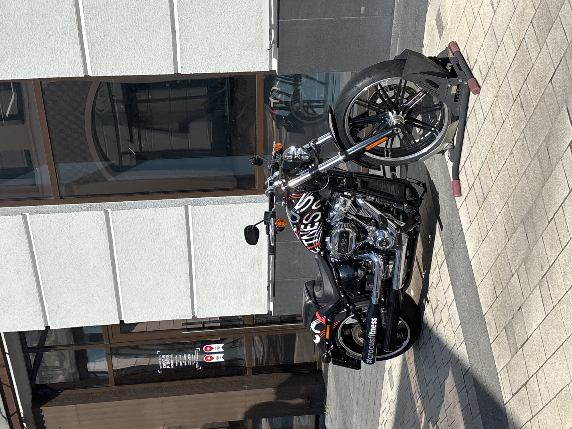 A black motorcycle with red and white graphics is parked on a sidewalk in front of a building. The motorcycle has a black seat and chrome accents. The building appears to be a commercial establishment with large windows. A sign on the window of the building displays text in Russian and the logo for "CROCUS FITNESS". The motorcycle is secured in a black stand. The sidewalk is paved with square tiles and illuminated by sunlight, casting shadows. The location is identified as Millennium Park, Russia.
