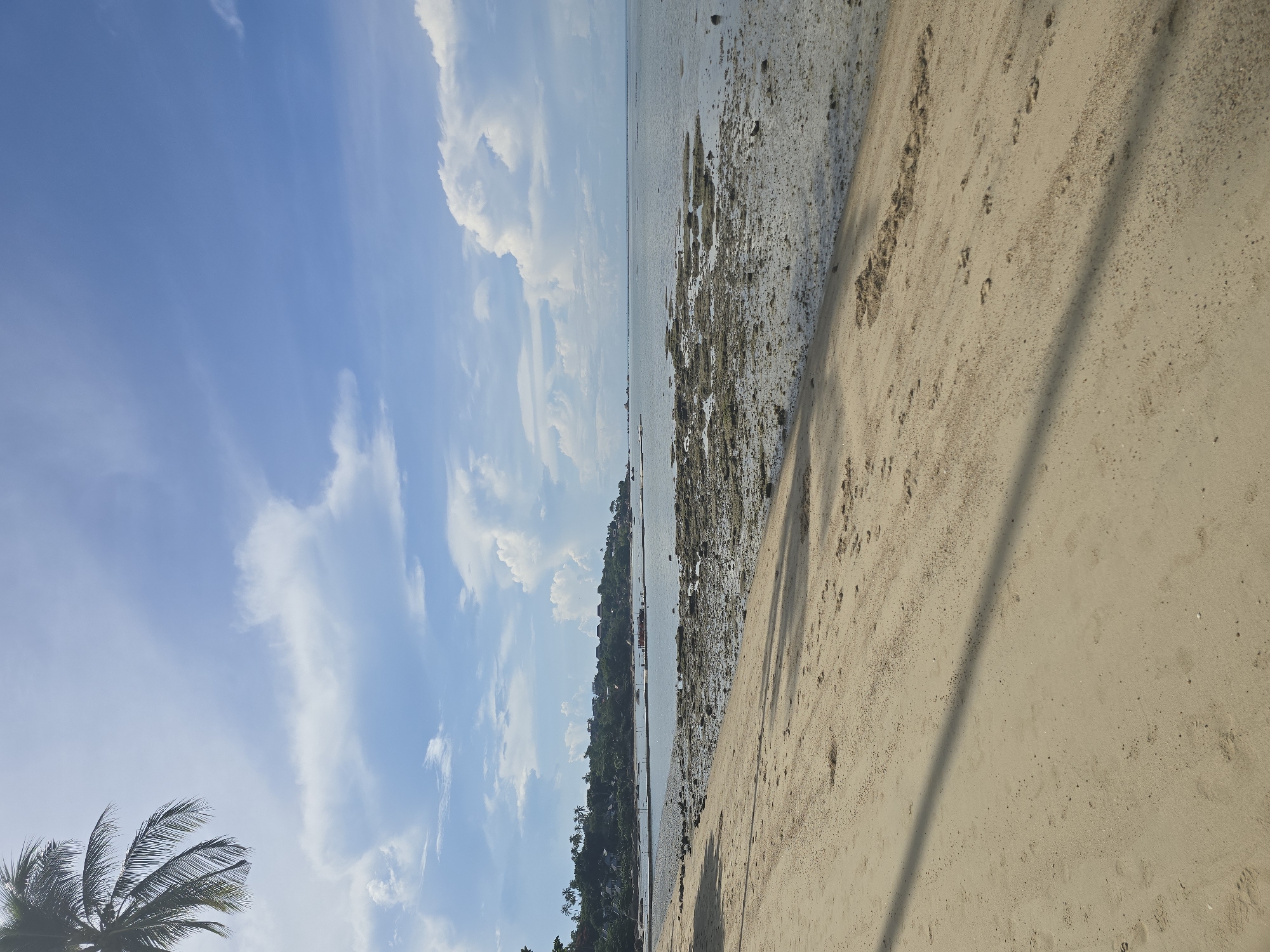 The image shows a beach scene in Baan Chaweng Noi, Thailand. In the foreground, a sandy beach is visible, with some footprints and shadows cast by palm fronds. The midground features a shallow body of water with exposed rocks and sandbanks, indicating a low tide. In the distance, a tree-covered hillside with buildings is visible, suggesting a coastal settlement. The sky above is blue with scattered white clouds, and a partial view of a palm tree is present in the upper left corner of the image. The overall scene depicts a serene tropical beach environment.