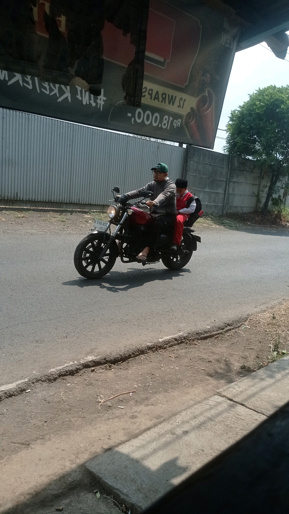 Two individuals, an adult male and a child, are riding a red motorcycle on a paved road in Bedali, Indonesia. The adult male, wearing a green cap, glasses, and a grey long-sleeved shirt, is operating the motorcycle. The child, wearing a red shirt and white pants, is seated behind him. The motorcycle has a license plate that reads "N 5760 AGT". The road is bordered by a dirt shoulder with some sparse vegetation. In the background, a grey corrugated metal fence runs along the side of the road, and behind that, a concrete wall and trees are visible. A large, weathered advertisement banner hangs above the fence, with partially visible text including "#thi KKIEKI" and "rp 18.000.-". The sky is bright, suggesting daylight.