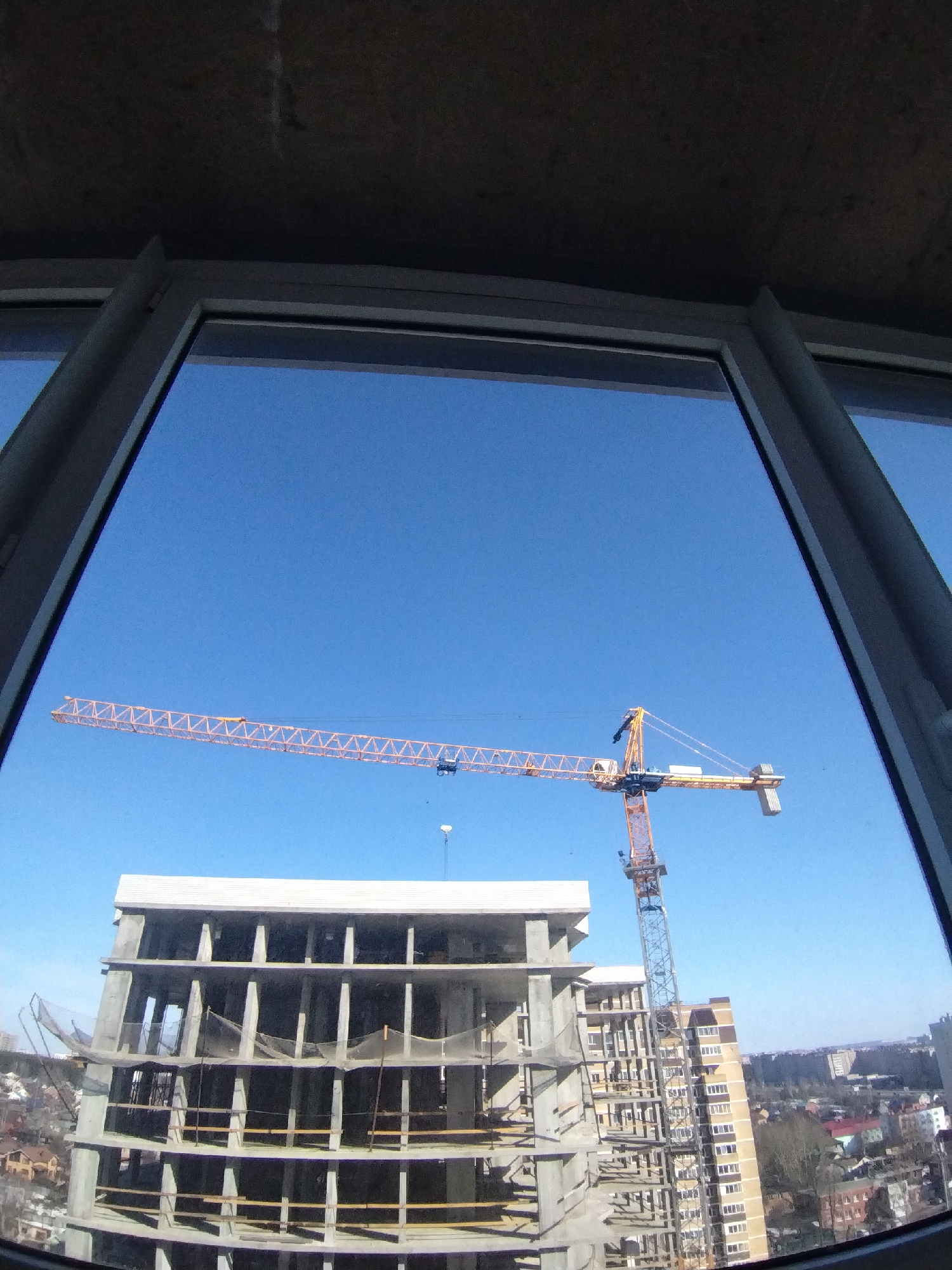 The image shows a construction site in Izhevsk, Russia, seen through a window. A tall crane dominates the scene, positioned next to a partially constructed building with exposed concrete beams and unfinished walls. In the background, there is a cityscape with residential buildings, trees, and other structures. The sky is clear and blue.