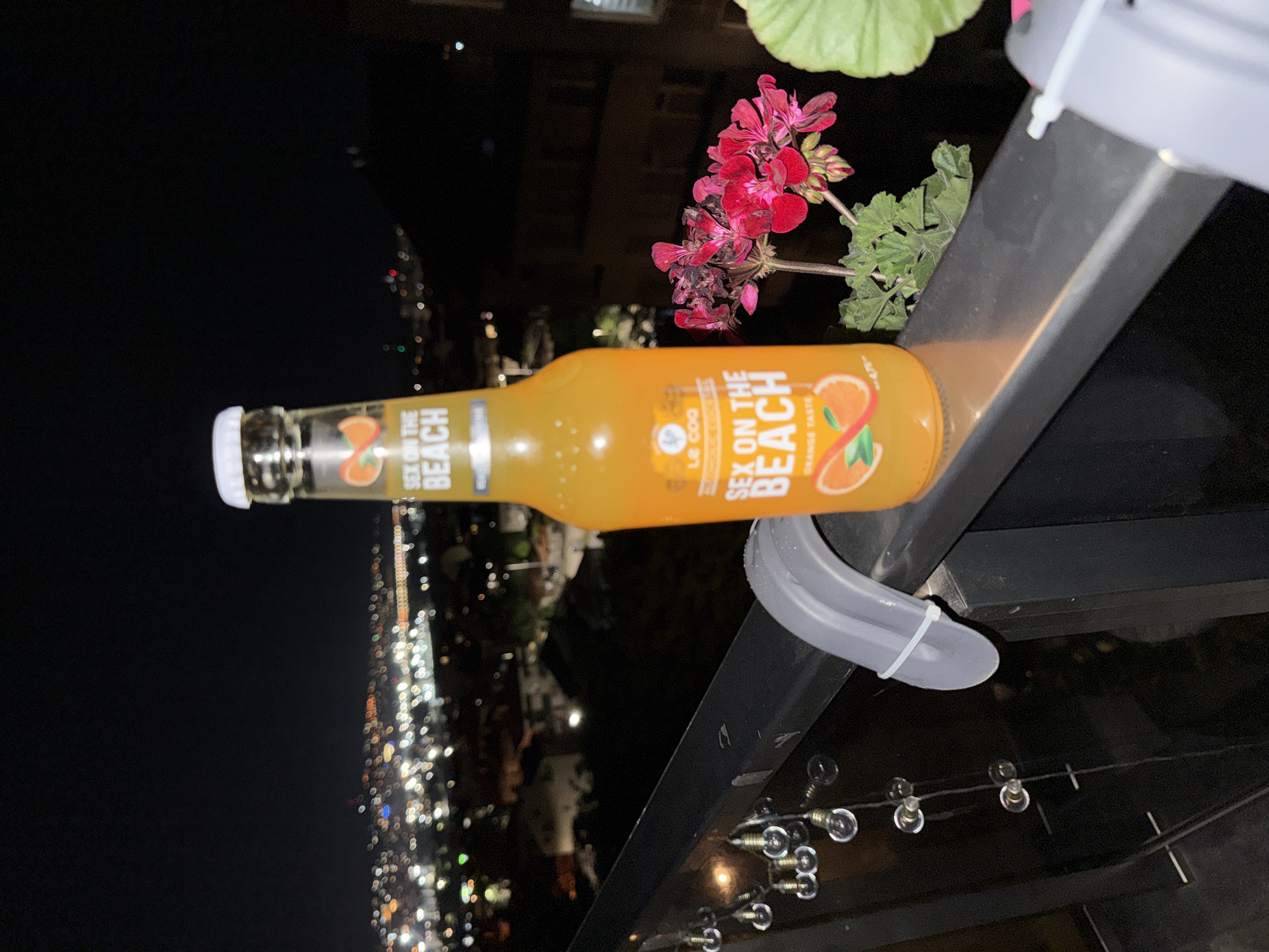 In an outdoor setting at night, with a cityscape visible in the background, a bottle of "Sex on the Beach" orange-flavored drink is placed on a railing. The bottle is clear glass with an orange liquid. The label features text and an illustration of an orange slice. Next to the bottle are pink flowers and green foliage. A string of small, clear lightbulbs is attached to the railing to the left of the bottle. A gray plastic object with a white zip tie is also attached to the railing. The overall scene appears to be on a balcony or terrace overlooking a city at night.