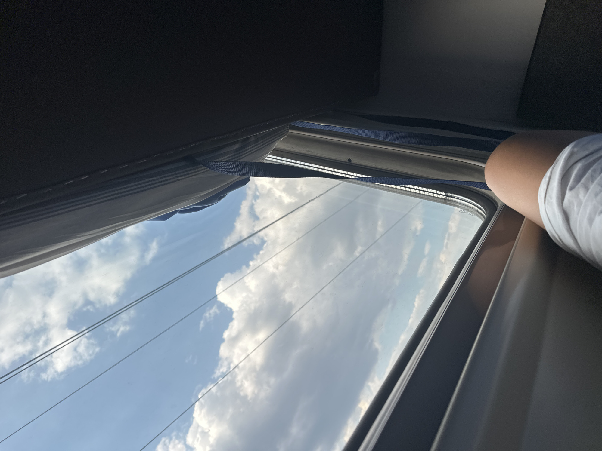 An image taken from the interior of a train shows a view out of a window. The sky is visible with white, fluffy clouds against a blue background. Several thin, dark lines, possibly wires, are superimposed over the sky. On the right side of the image, a dark, upholstered surface, likely part of the train's interior, takes up a significant portion of the frame. A blue strap hangs down from this surface. In the foreground, on the lower left, a person's bare leg wearing white shorts is visible. The scene appears to be in Krasiukovskaya, Russia, as indicated by the prompt.