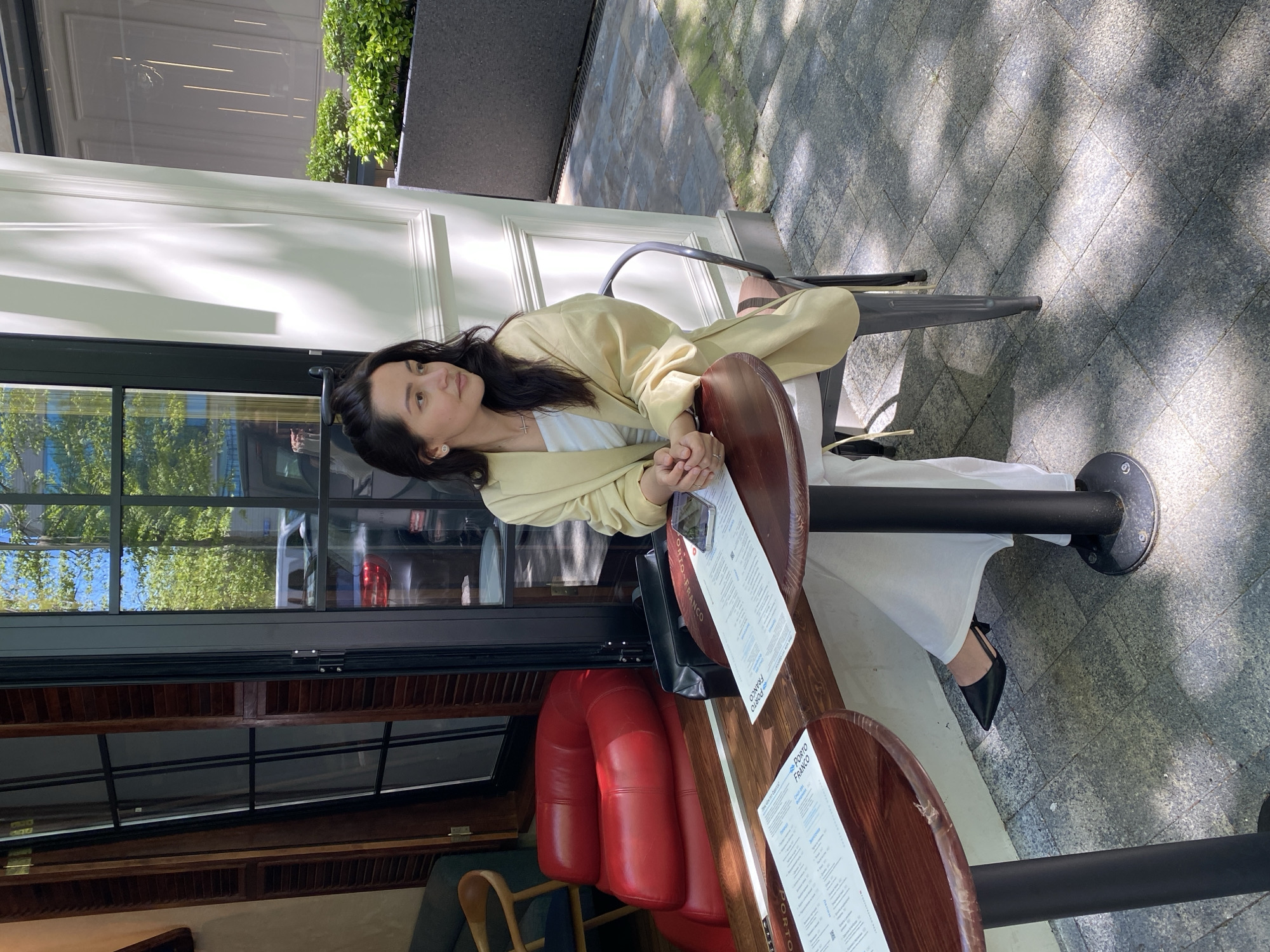 A woman with long, dark hair is seated at an outdoor cafe table in bright daylight. The image is rotated 90 degrees clockwise, depicting the scene with the ground to the right and the building to the left. She is wearing a light yellow blazer over a white top, white wide-leg pants, and black pointed-toe flat shoes. A small cross necklace and earrings are visible. Her hands are clasped on the dark wooden table, and she gazes calmly to her left.
The setting is an outdoor seating area of a cafe or restaurant, likely in Sochi, Russia. The woman is at one of two dark wooden tables, each with a black metal base. Menus on both tables prominently display the text "PORTO FRANCO" at the top. She is seated on a black metal chair with a wooden back. Behind her, a modern building facade features white paneled walls and large windows that reflect trees and the sky. Through the windows, glimpses of red upholstered seating can be seen inside the establishment. The ground is paved with grey interlocking tiles, dappled with strong sunlight and shadows, indicating a clear, sunny day. Green foliage is visible in the background, suggesting planters or landscaping. The scene conveys a relaxed moment at an urban outdoor dining spot.