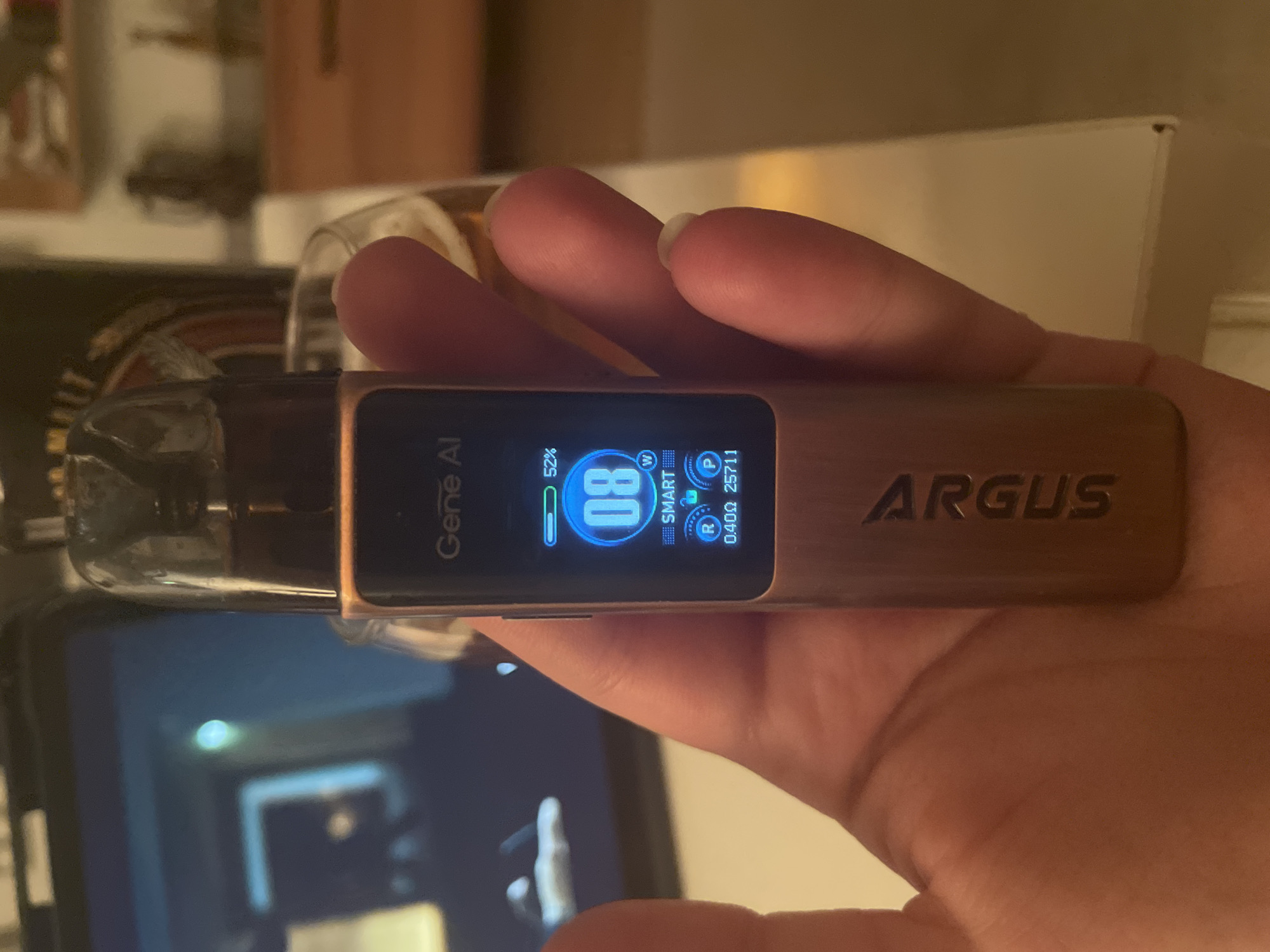 The image shows a close-up of a person's hand holding a vape device. The vape device has a digital screen displaying "Gene AI," a battery indicator at 52%, a wattage of 8W, and "SMART." Below this, there are icons labeled "R" and "P" with corresponding numbers. The device itself is rose gold colored and has "ARGUS" written vertically on its side. In the background, a tablet screen is visible, showing a blurred image. To the right of the vape, a glass with a beverage is partially visible, along with shelves in the background. The setting appears to be indoors, possibly a home or a bar, with warm lighting. There is no specific indication of the location being in Yunus Emre, Turkey.