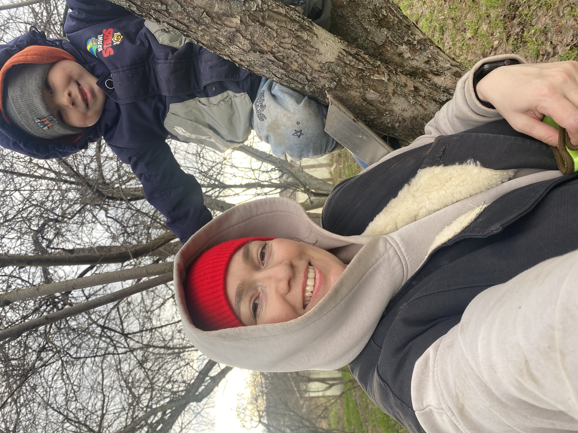 A candid outdoor photograph taken in Ольховка, Russia, features a woman and a child smiling at the camera. The image is oriented sideways, with a large tree trunk running diagonally through the frame.
In the foreground, to the right, a woman smiles broadly, looking towards the viewer. She wears a red beanie under a light beige hooded sweatshirt with the hood up, and a dark jacket with a visible cream-colored sherpa lining. A black smartwatch is visible on her left wrist, and she holds a green and yellow object in her hand.
Above and to the left of the woman, a young child also smiles, looking at the camera. He is dressed in a navy blue winter jacket with grey sections, an orange hood, and a grey beanie. The jacket has "BRAWL STARS" and star logos visible on the chest. Text reading "STARS" with a star icon is also visible on the child's light-colored jeans.
The setting appears to be a natural area, likely a park or wooded space, characterized by the prominent tree trunk and numerous bare deciduous branches against a bright, possibly overcast, sky. Green grass is visible on the ground in the background. Both subjects' warm clothing suggests cool weather, likely late autumn, winter, or early spring. The expressions of happiness on both faces indicate a joyful outdoor activity.