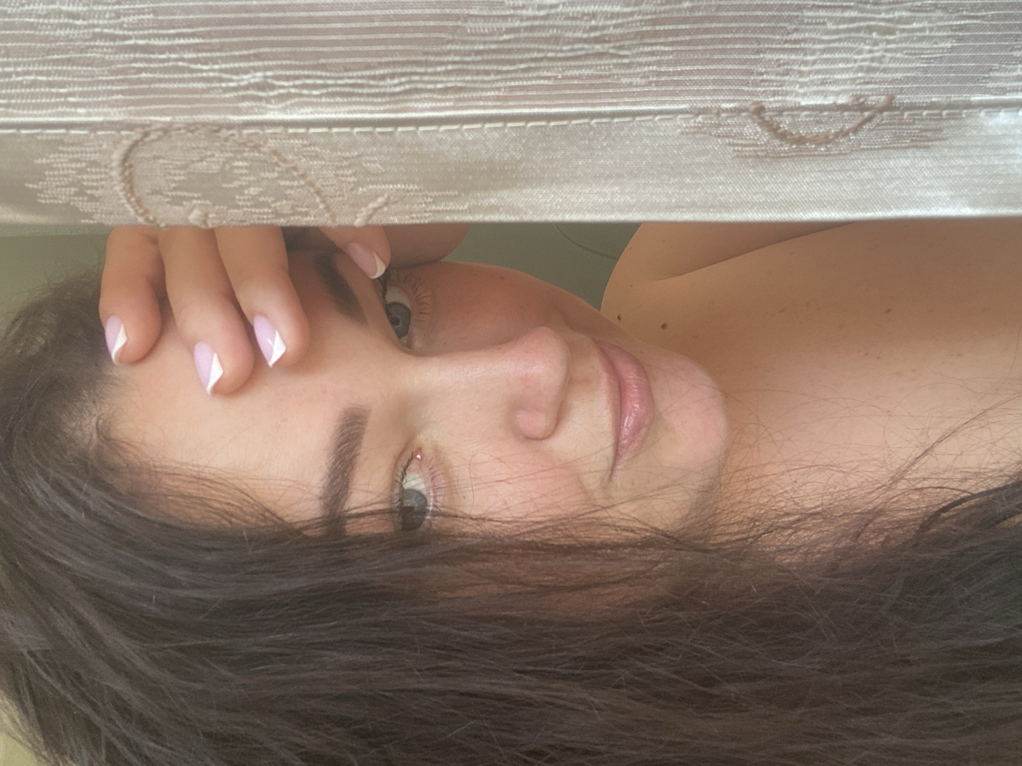 A woman with brown hair and fair skin is partially visible, lying down. The woman's face is partially obscured by her hand. Her nails are manicured with a white French tip. A patterned, light-colored fabric is directly above her head, suggesting she is in an interior space. The image does not contain sufficient information to identify the location in Sochi, Russia.