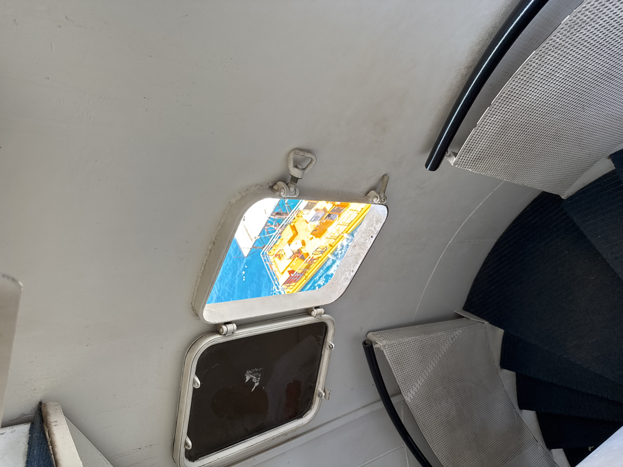 An interior view looking down a spiral staircase towards a porthole window that looks out onto bright blue water and a yellow boat. The staircase has dark, carpeted steps and white meshed railings. The walls of the interior are painted white. The porthole window is open and a metal latch is visible. The boat appears to be a pleasure craft or a work boat. The scene is likely within a lighthouse or a similar structure along the coast of Eilat, Israel, given the clear water and the type of boat visible.