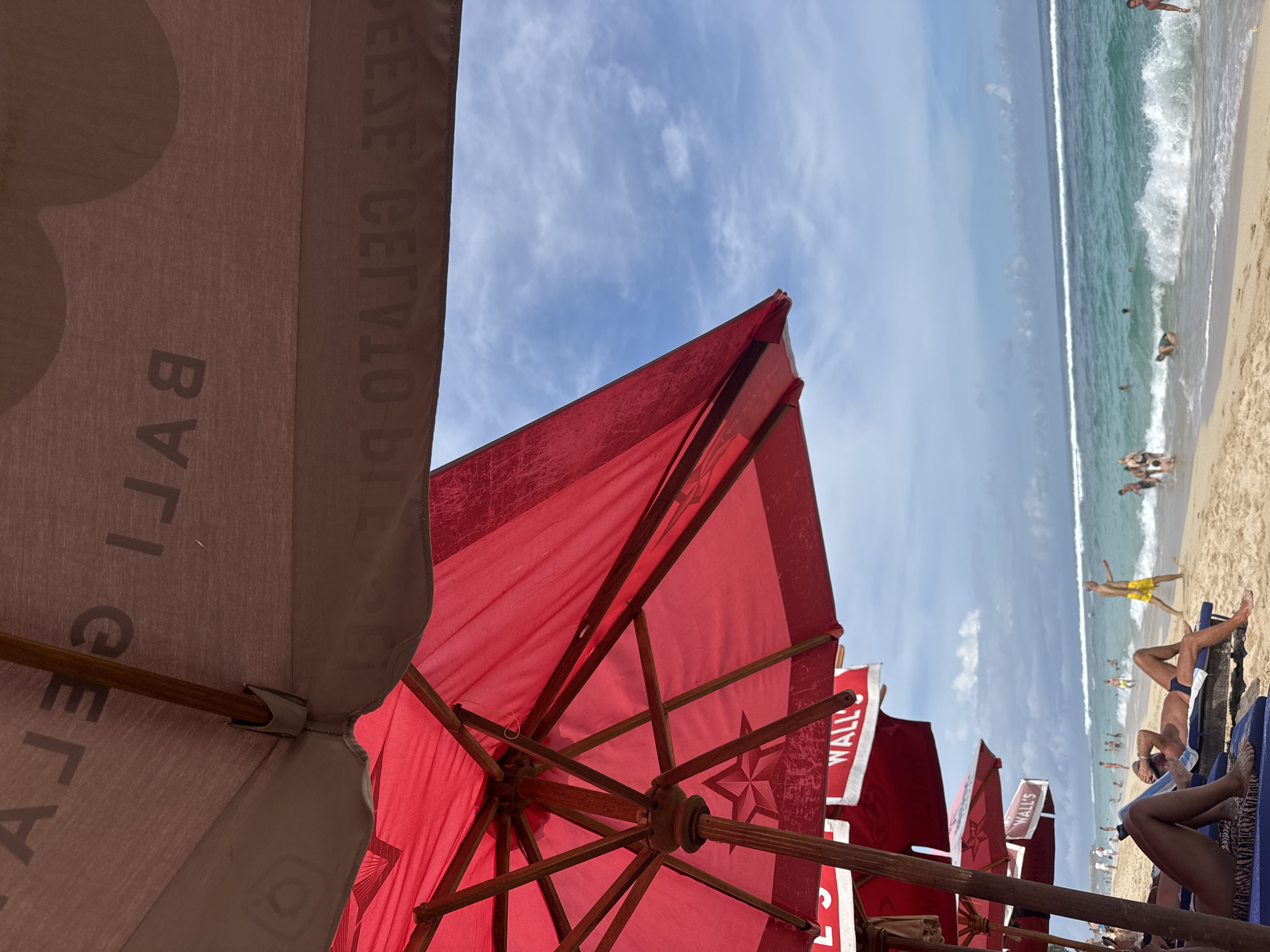 This image shows a beach scene, most likely in Kuta Selatan, Indonesia, given the "Bali Gelato" logo on an umbrella in the foreground. There are several red umbrellas lining the beach, interspersed with occasional beige umbrellas. The red umbrellas appear to be branded with "WALL'S" in white lettering. Beachgoers are visible in the background, engaging in various activities. The ocean is visible in the background as well. The sky is blue with scattered clouds.