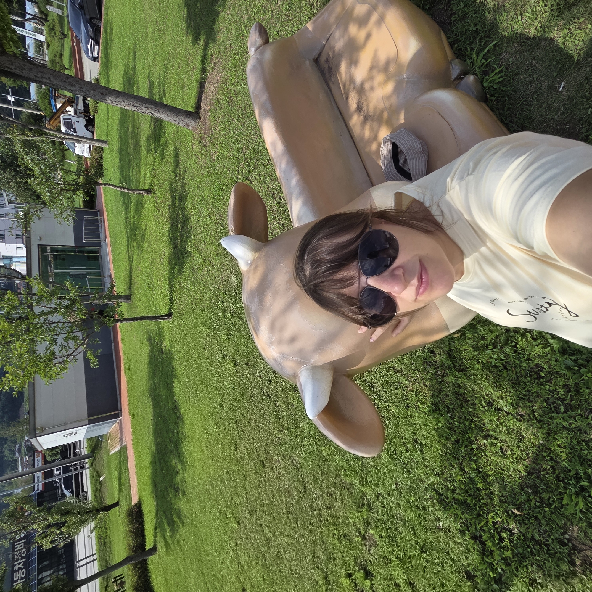 A woman wearing sunglasses and a light-colored t-shirt takes a selfie outdoors, sitting next to a large, tan, stylized sculpture of a reclining deer. The sculpture is large enough to sit on and has horns and ears. A striped hat rests on the sculpture's back. The background shows a grassy area with trees, a paved walkway, and a building with a glass entrance. In the distance, there is a road with vehicles, including a white truck and a black car, and more buildings. The scene is set in Dunnae-myeon, South Korea, as indicated by signage in Korean. The lighting suggests it is daytime.