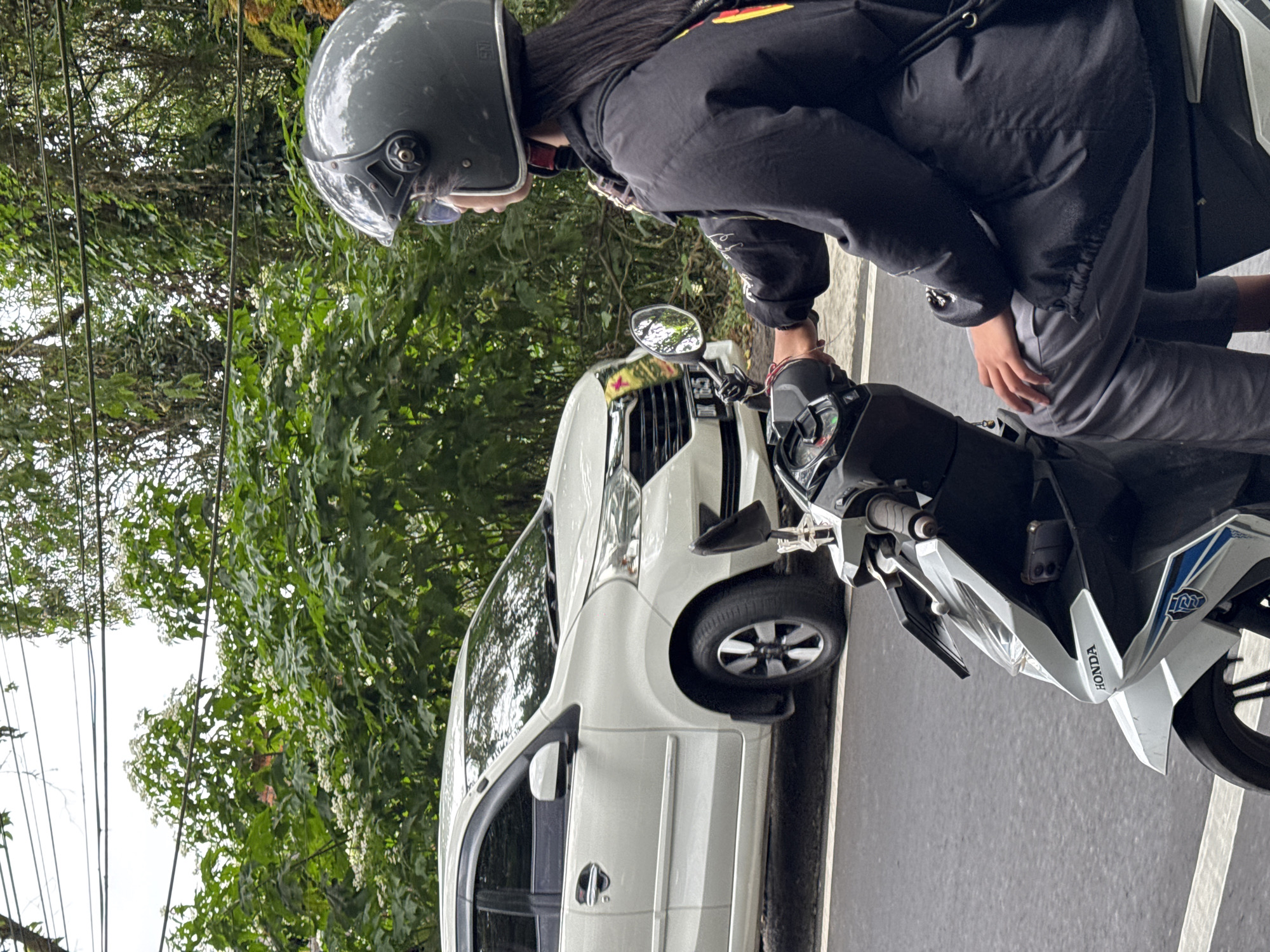 In Kintamani, Indonesia, a person wearing a gray helmet and a black jacket rides a white Honda scooter on an asphalt road. The rider's gray pants are visible. A white car is parked next to the road, with its front license plate reading "DK 165 M". Lush green trees and foliage line the background of the scene, with power lines overhead. The sky is overcast.