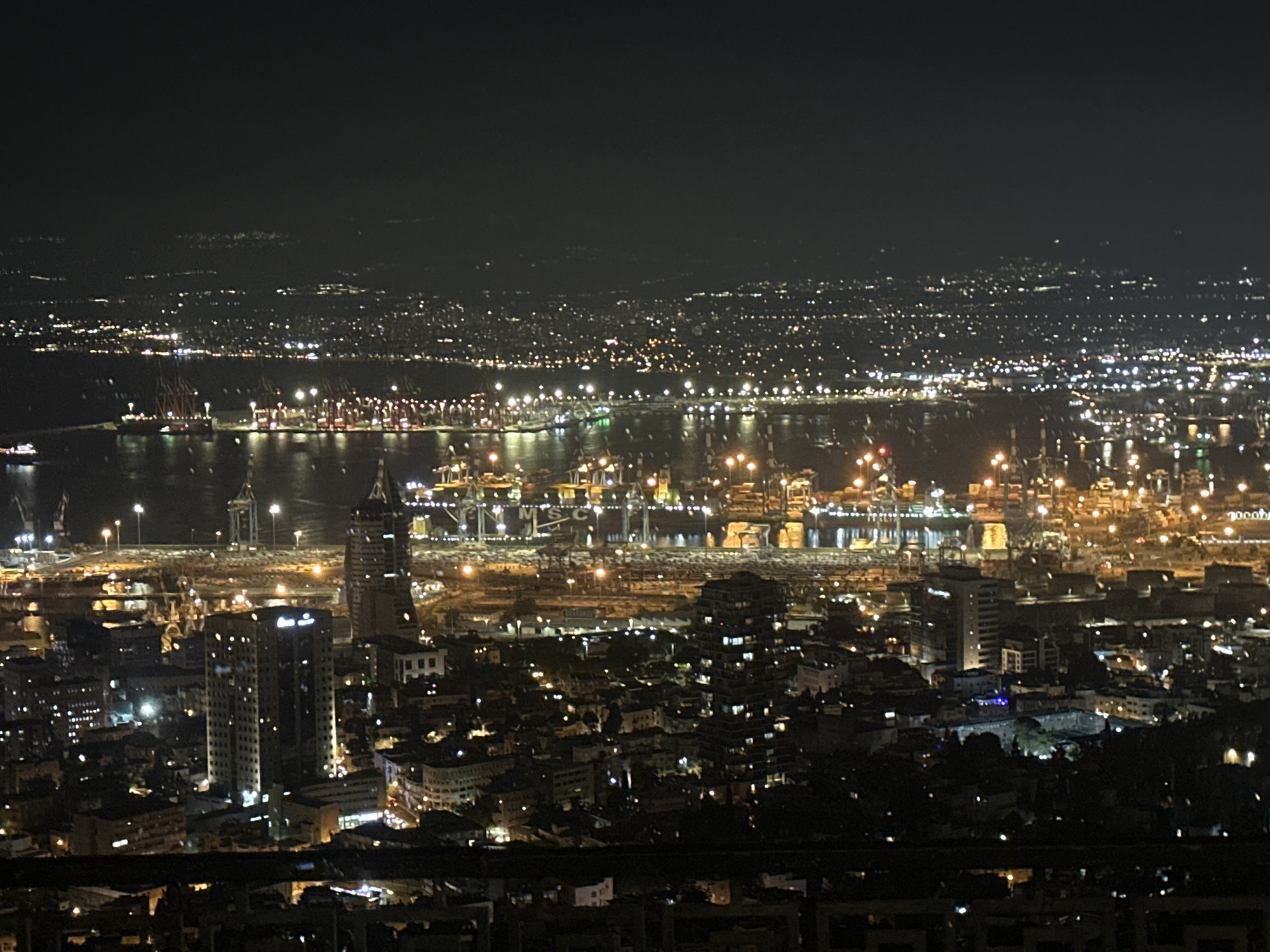 This is a high-angle, nighttime panoramic view of the city of Haifa, Israel, likely taken from Mount Carmel. The foreground shows a dark, blurred silhouette of railings or balconies, suggesting the vantage point is from a building or observation deck.
The midground is dominated by the illuminated cityscape of Haifa. Several distinct structures are visible:
- A prominent, tall, rectangular skyscraper with many lit windows is located centrally in the midground. It appears to be an office building or residential tower.
- To its left, a shorter, boxier building with visible windows is present.
- Further left, the silhouette of a tall, spire-like structure, possibly a communication tower or part of a larger complex, is discernible against the night sky.
- A large port complex stretches across the background, characterized by numerous bright lights, cranes, and ships. The industrial lights create a dense network of illumination.
- The water of the bay is visible, reflecting the lights of the city and the port, creating a shimmering effect.
- Beyond the immediate urban and port areas, a sprawling expanse of city lights extends into the distance, indicating the wider urban area of Haifa and potentially surrounding settlements. The distant lights are more diffused and less distinct due to the distance.
The environment is dark, with the primary light sources being artificial city and port illumination. The sky is black, with no visible celestial bodies or moon. The overall impression is one of a vibrant, active coastal city at night. No people are directly visible in the frame, and the focus is on the urban landscape and its infrastructure. The perspective suggests a wide view encompassing both the residential and industrial heart of Haifa.