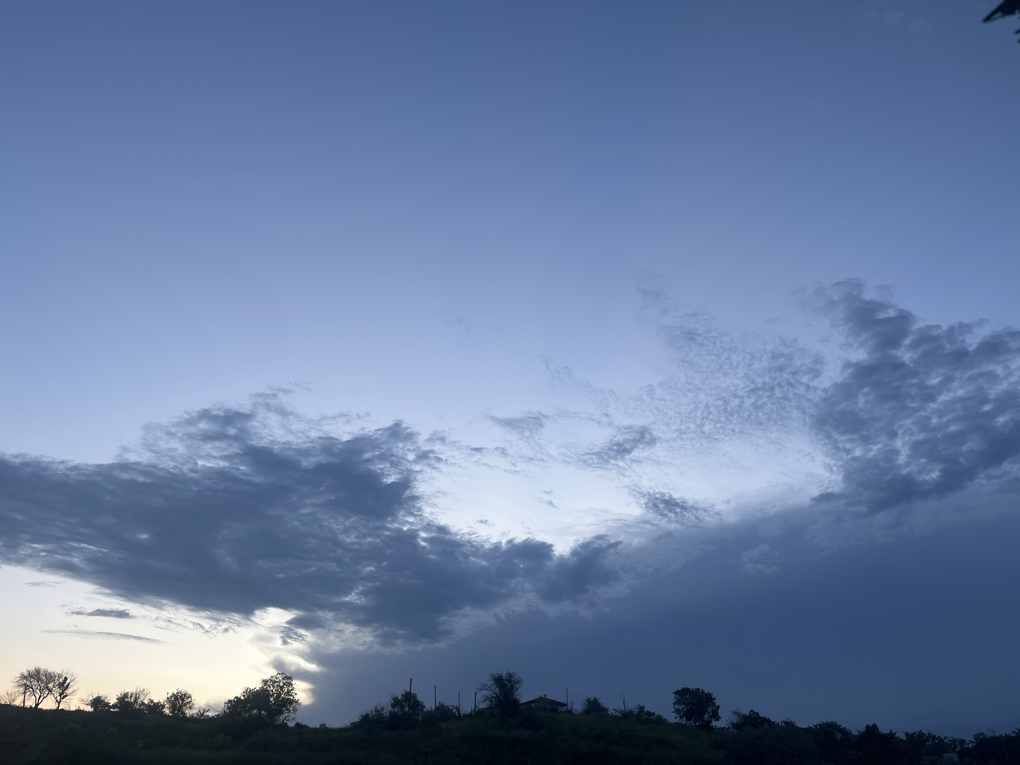 The image depicts a twilight sky over a rural or semi-rural landscape, situated in the vicinity of Varna, Bulgaria. The scene captures either dawn or dusk, characterized by a transition of natural light.
The upper three-quarters of the frame are dominated by the sky. The uppermost section displays a clear, pale desaturated blue. Below this, a complex system of clouds extends across the middle and lower sky. On the left, denser, darker stratocumulus clouds are present, with a faint, pale yellow-orange glow emanating from the horizon directly behind them, indicating the position of the sun. Towards the center and right, the cloud formations become more intricate, featuring patches of altocumulus and developing cumulus clouds, varying in texture and density. A significant mass of dark, heavy cloud occupies the lower right section, descending towards the horizon, suggesting a localized weather system or rain.
The lower quarter of the image shows a dark, silhouetted landscape, consistent with a series of hills or elevated terrain. Numerous trees, some appearing bare and others with denser foliage, are discernible along the crest of the hills. A single-story structure, likely a house, with a visible reddish-brown roof, is located on the hilltop slightly to the right of the center. Several utility poles or similar slender vertical structures are also present on the ridgeline near the house and elsewhere. The landscape is presented as an almost complete silhouette against the brighter sky. No people, animals, or moving vehicles are visible within the frame. The overall atmosphere conveys a calm yet dramatic natural light event.