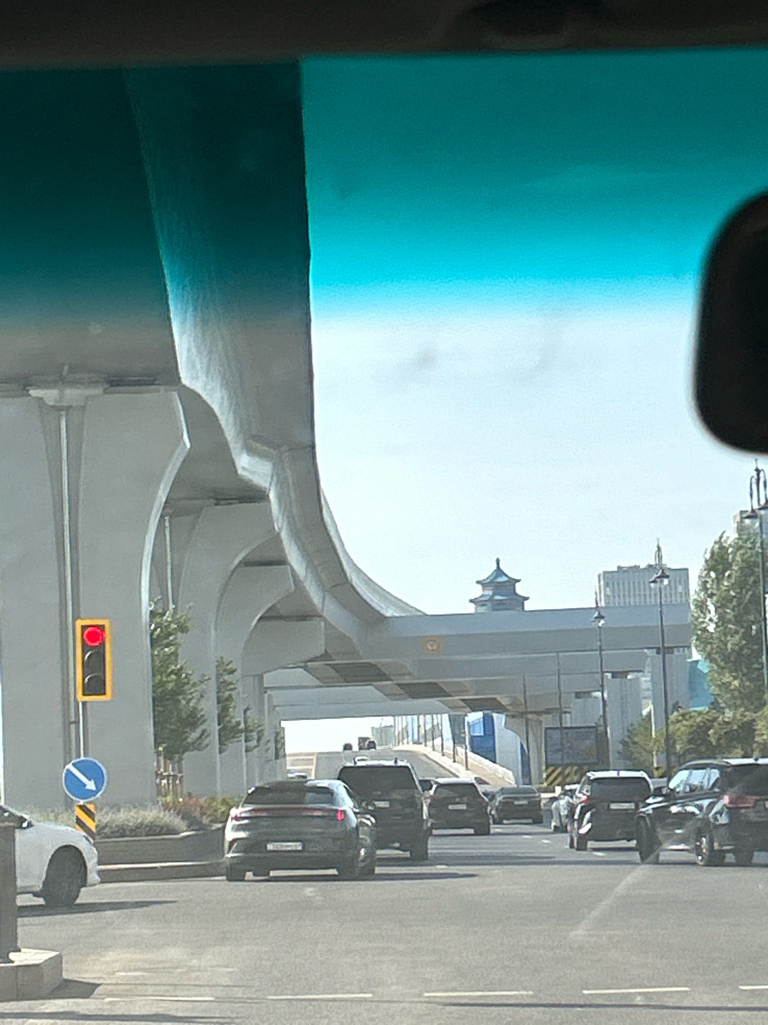 The image captures a street scene in Astana, Kazakhstan, with traffic moving under a large elevated road structure. In the foreground, several vehicles are visible, including a white car on the left and a dark SUV in the center. A traffic light displaying a red signal is prominent on the left side of the road. Behind the immediate traffic, more cars are seen, and the elevated road arches overhead, supported by large concrete pillars. In the distance, a building with a distinctive tiered roof, reminiscent of traditional East Asian architecture, stands out against the sky. The overall environment suggests an urban setting with significant infrastructure.