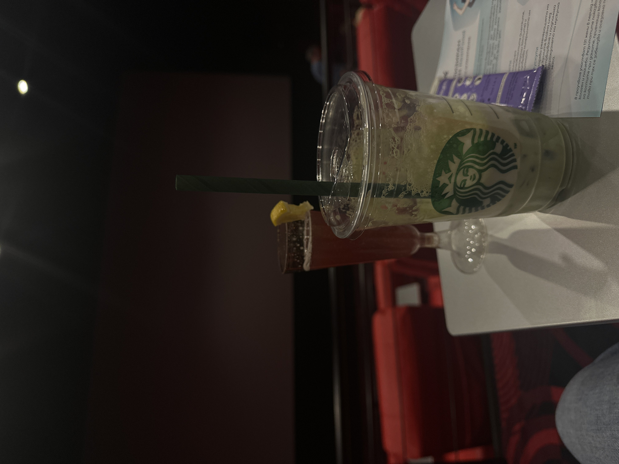 The image captures a dimly lit indoor scene, likely a cinema or theater, taken from a slightly elevated perspective. In the foreground, two beverages rest on a small, white table. To the right, a partially consumed Starbucks plastic cup, containing a light green or yellowish drink with a green straw, is prominently displayed with its iconic siren logo. To its left, a stemmed glass holds a reddish liquid, garnished with a lemon wedge. A piece of paper with visible text is also on the table, partially obscured by the Starbucks cup.
The background is very dark, with muted red seating visible, characteristic of a movie theater or performance hall. The dim lighting suggests an ongoing show or movie. A bright, out-of-focus light source appears in the upper left corner of the frame. A person's leg, wearing blue jeans, is partially visible in the lower right, indicating a seated viewer.
The visible text on the paper is in Bulgarian and appears to be an informational or promotional leaflet, mentioning terms like "мускули" (muscles), "колаген" (collagen), and "регенерацията" (regeneration), suggesting content related to health or supplements. The overall atmosphere is one of leisure and relaxation during an evening entertainment event in Sofia, Bulgaria.