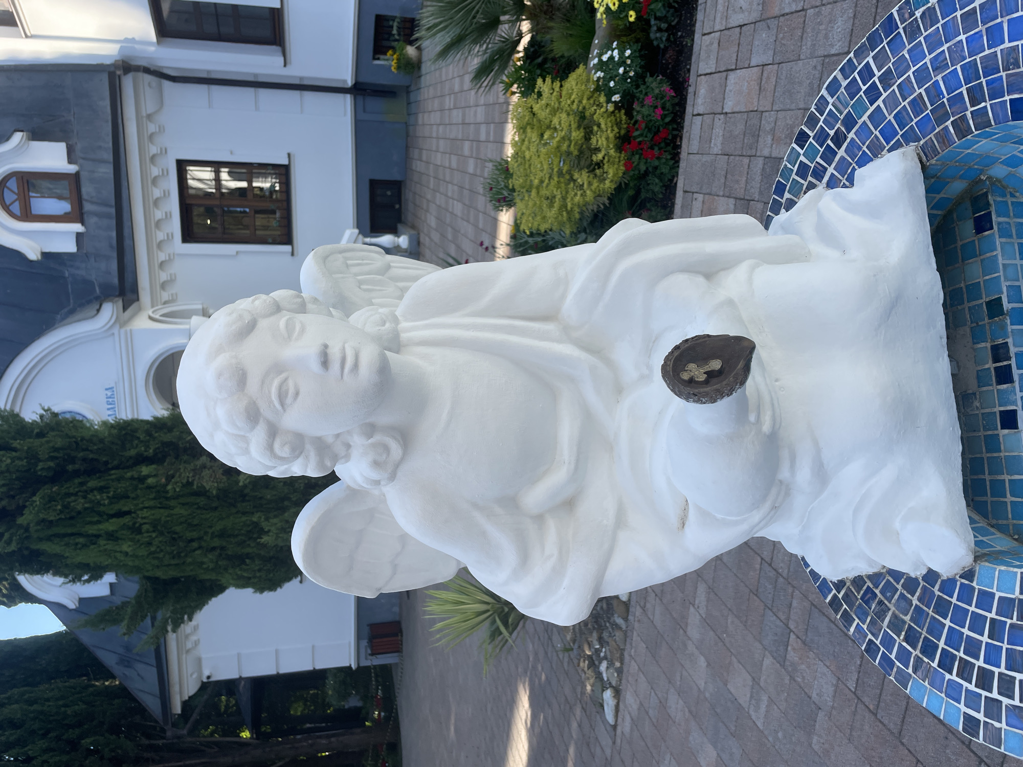 The original image, which appears to be rotated 90 degrees clockwise, presents a serene outdoor scene centered around a white angel statue.
In the foreground, a detailed white statue of an angel, possibly a cherub or putto, is prominently featured. The angel has soft, curly hair, wings, and a serene expression on its face. It appears to be sitting or kneeling, holding a small, dark, circular object adorned with a golden cross-like symbol in its hands. The statue rests on a patterned paved surface, composed of light-colored interlocking blocks, and is situated next to a curved structure covered in various shades of blue mosaic tiles, resembling part of a fountain or decorative basin.
The background reveals a well-maintained garden with lush greenery, including tall evergreen trees, palm fronds, and vibrant flowering bushes with red, white, and yellow blossoms. Beyond the garden, several buildings with distinct architectural styles are visible. These buildings feature white facades and dark grey or blue roofs. They have a mix of arched and rectangular windows, some with dark wooden frames, and exhibit decorative architectural elements. On one of the prominent white buildings, the Cyrillic text "ЛАВКА" (meaning "SHOP" or "STORE") is visible above an archway.
The scene is illuminated by bright natural daylight, indicating clear, sunny weather. Soft shadows suggest either mid-morning or late afternoon. The overall setting is tranquil and decorative, hinting at a landscaped public space or private garden in a location like Sochi, Russia, as suggested by the visible text and architectural style. No specific activity or event is taking place; the image captures a static, picturesque moment.