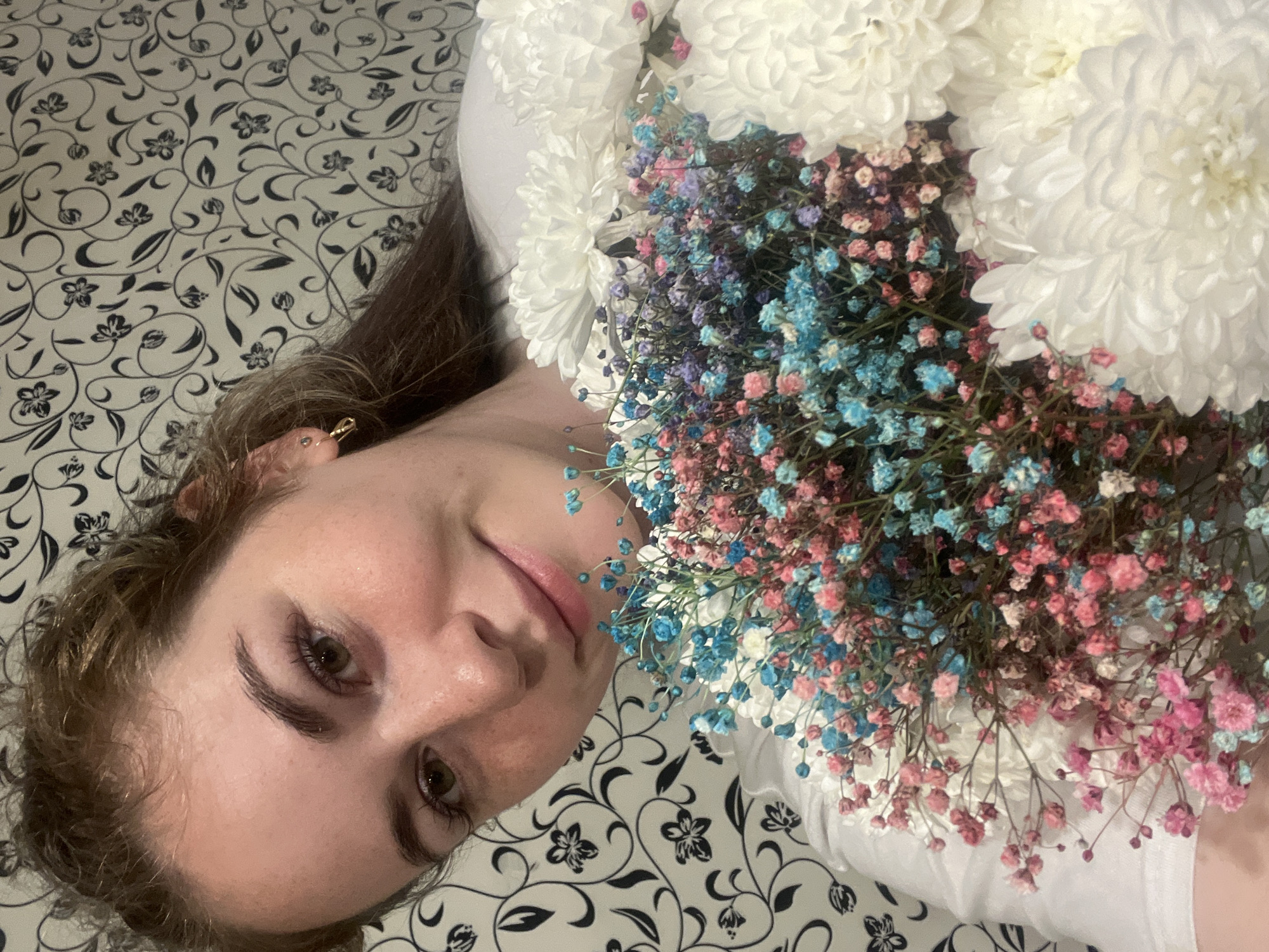 A woman with fair skin and brown hair is lying on a bed with a patterned black and white bedspread. She is holding a bouquet of white chrysanthemums and multicolored baby's breath flowers. The flowers partially obscure her upper body. She is wearing a white shirt. She is looking directly at the camera. The location cannot be determined from the image.