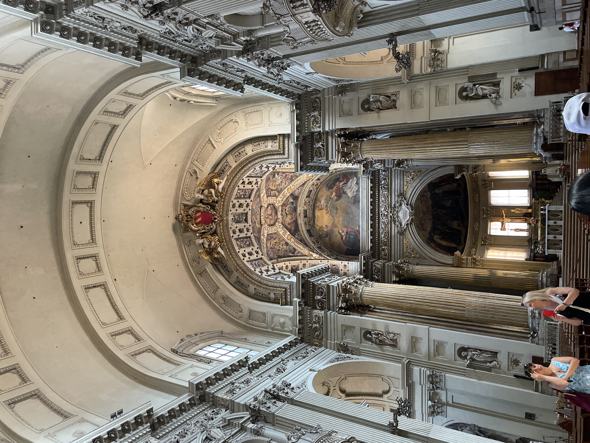 The image captures the ornate interior of a church in Bologna, Italy, showcasing its architecture. The ceiling is elaborately decorated with paintings, sculptures, and architectural details. Massive columns line the sides, adorned with statues. Several people are visible in the image, including two women in the foreground. One of the women is wearing a blue dress, and the other has blonde hair and is wearing a black top. They are near the pews of the church. Other individuals are blurred and partially visible in the background, suggesting a small gathering. The image is taken from an upward angle, emphasizing the height and grandeur of the church's interior.