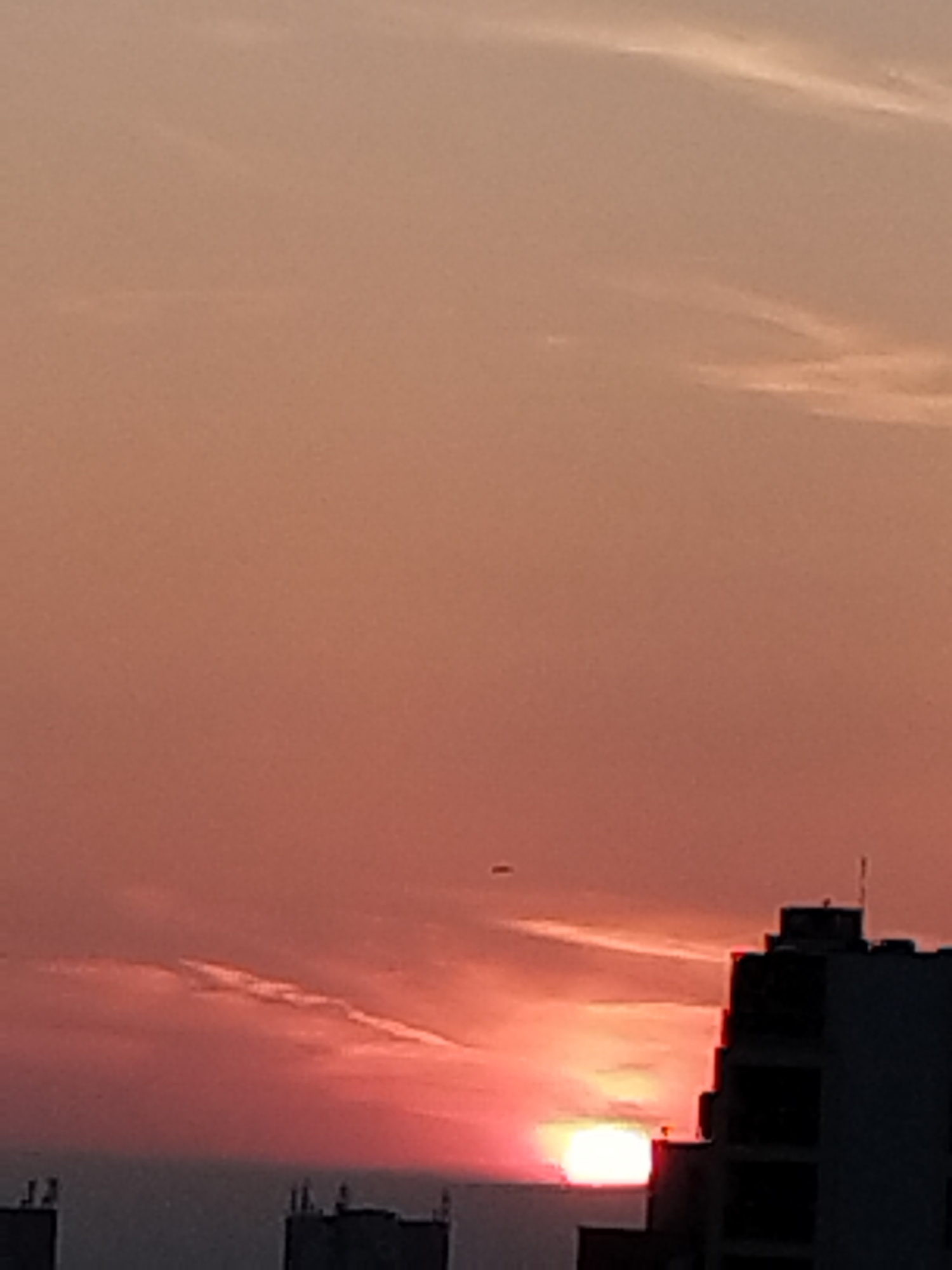 The image depicts a sunrise or sunset over an urban landscape in Unknown, Unknown. The sky presents a vivid gradient of warm colors, transitioning from a muted orange-brown in the upper portion to deep reds and vibrant pinks closer to the horizon. A prominent, elongated cloud streak, illuminated from below, extends horizontally across the lower-mid sky. The sun, a bright white-yellow orb, is partially visible at the horizon on the right, emitting an intense glow. A small, dark, oblong object is present in the upper-middle-left area of the sky. In the foreground and midground, multiple buildings are silhouetted against the bright sky. A tall, multi-story building with visible architectural steps and roof antennae occupies the right side of the frame, partially obscuring the sun. Two smaller, dark building structures with roof protrusions are visible in the lower-left. No individuals or explicit actions are depicted.