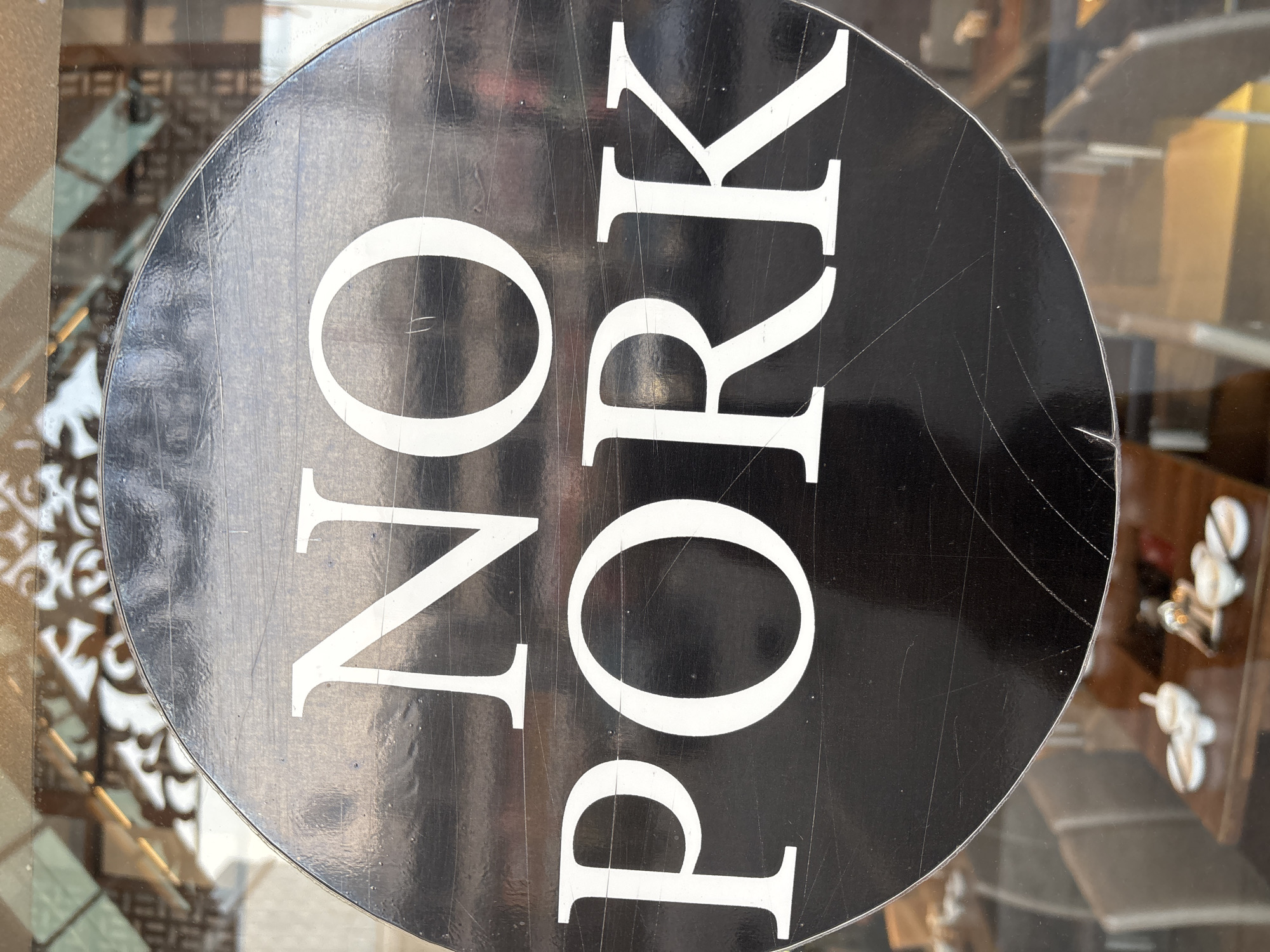 A circular sign with white text reading "NO PORK" is affixed to a glass surface. The sign is black with white serif font. The background behind the sign is blurred, showing what appears to be an interior space with tables and chairs. There are reflections on the glass surface, including light and some decorative architectural elements. The location of Pekanbaru, Indonesia, is not discernible from the image itself.