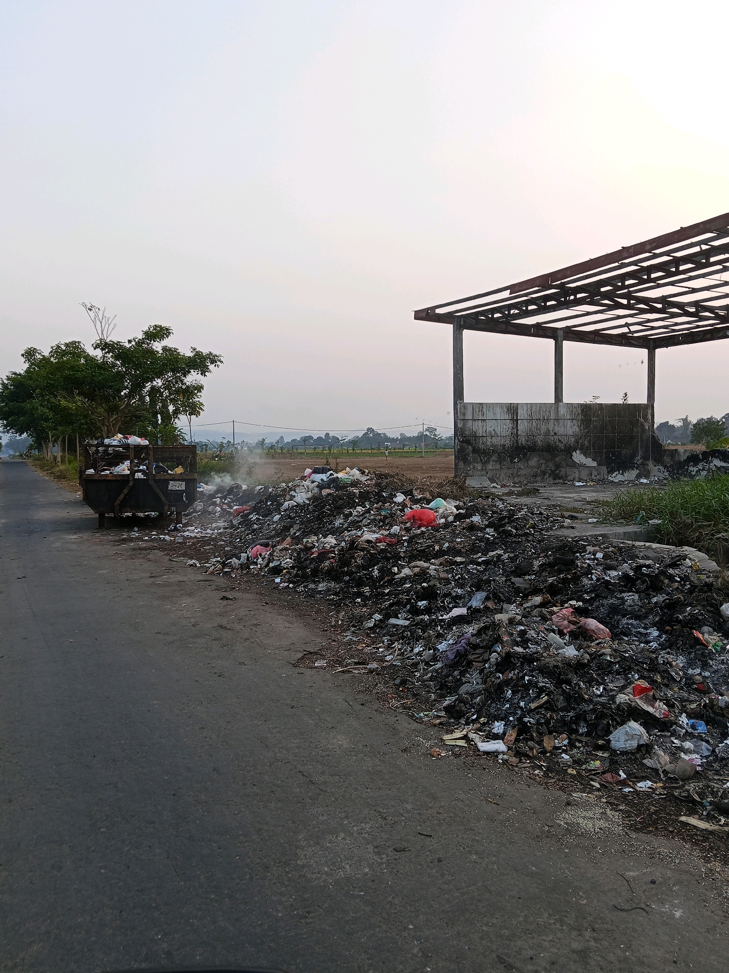 The image depicts an outdoor scene in Gaprang, Indonesia, characterized by significant environmental pollution. A large, overflowing dumpster filled with assorted waste is positioned on the left side of an asphalt road. Adjacent to the road, a sprawling pile of trash, consisting of various discarded materials and plastic bags, is accumulating. Some of the garbage appears to be smoldering, emitting a visible plume of smoke, indicating potential spontaneous combustion or deliberate burning. In the background, an unfinished or abandoned structure with a skeletal metal roof and concrete walls stands as a stark contrast to the surrounding natural landscape, which includes trees, fields, and a hazy sky. The overall impression is one of neglect and unsanitary conditions.