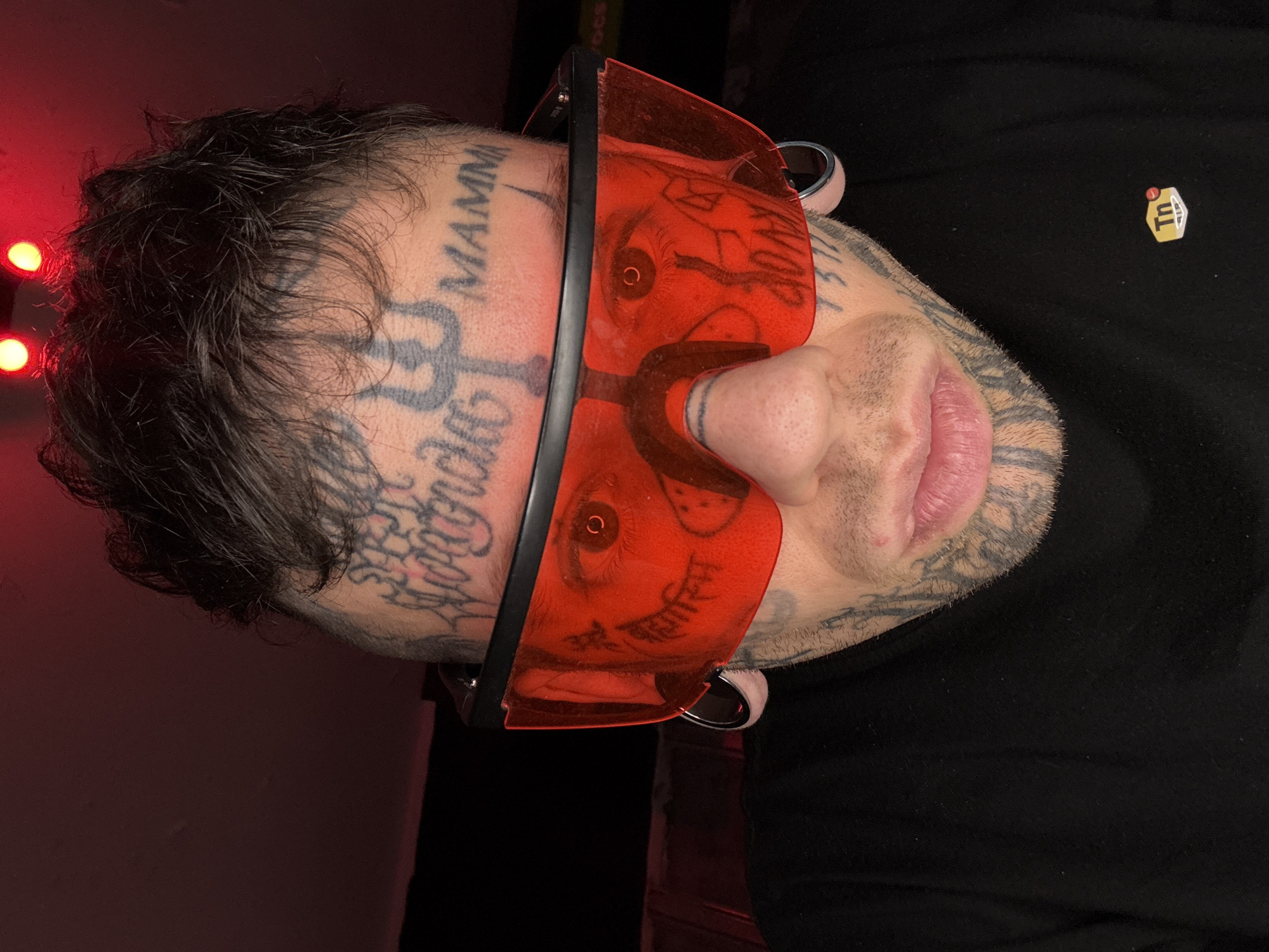 This close-up image depicts an adult individual situated in an indoor environment in Buenos Aires, Argentina. The subject, viewed from the head to the upper torso, presents with dark, dishevelled hair and extensive tattooing across the face and scalp. A prominent trident symbol is tattooed centrally on the forehead, with the word "MAMMA" visible on the right forehead and script lettering on the left forehead. Further script and symbolic tattoos are visible across the cheeks, jawline, and a horizontal line tattoo is present on the nasal bridge. The individual wears large, red-tinted, rimless-style sports sunglasses with black temples, through which the eyes are partially discernible. Both earlobes feature large-gauge metal tunnel piercings. The subject is dressed in a black long-sleeved garment, displaying a "Tn Air" logo on the left chest. The scene is illuminated predominantly by red light, with two distinct red light sources visible in the upper background against an otherwise dark and indistinct backdrop.