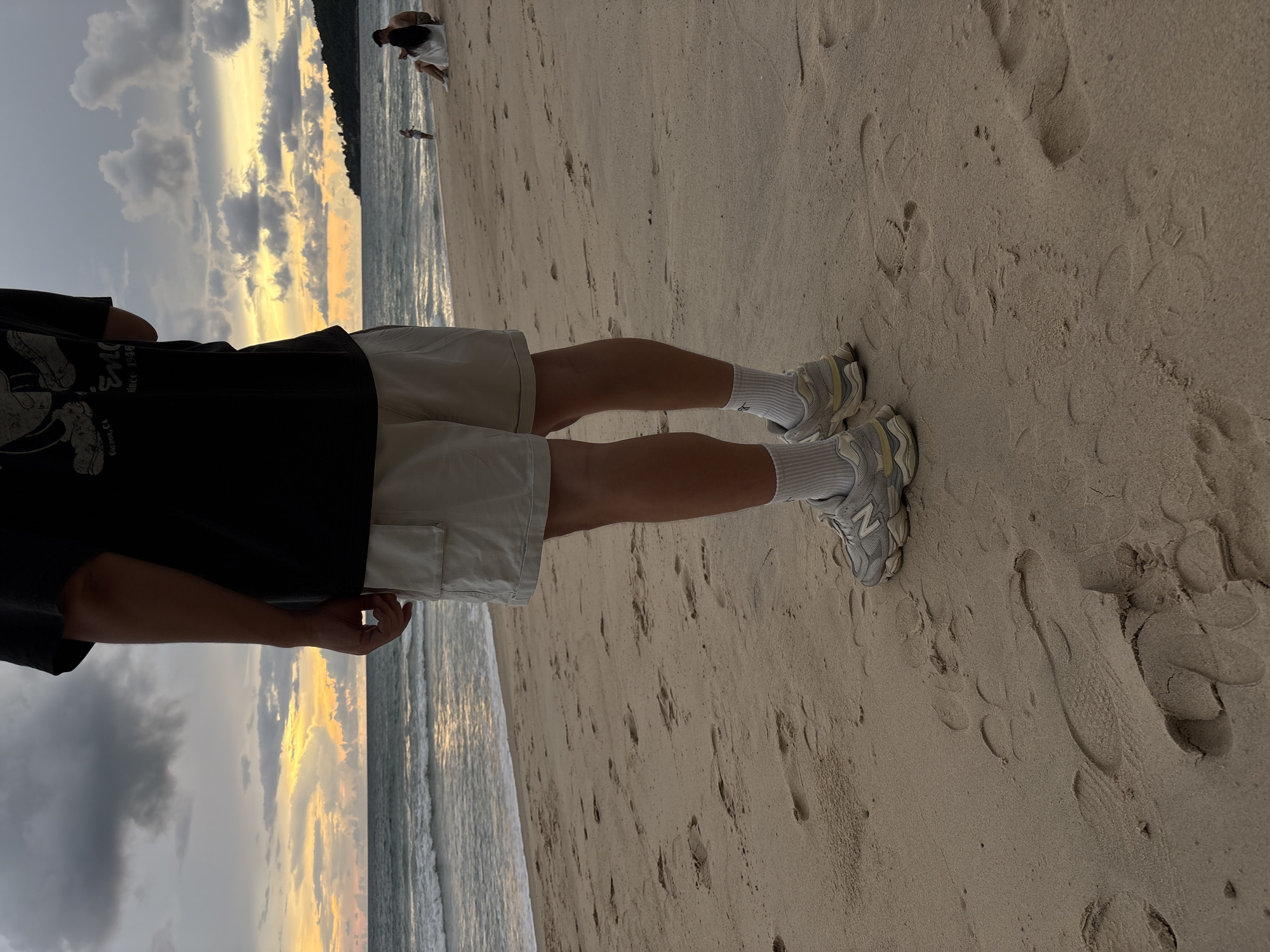The image shows the lower body of a person standing on a sandy beach in Choeng Thale, Thailand. The person is wearing a black t-shirt, light-colored shorts, white socks, and grey athletic shoes. The beach has many footprints in the sand. In the background, the ocean waves are visible, and the sky is cloudy with hints of orange and yellow from a sunset. Further in the distance, there are two people sitting on the beach, and another person standing near the water. A landmass with vegetation is visible on the right side of the frame, against the horizon.