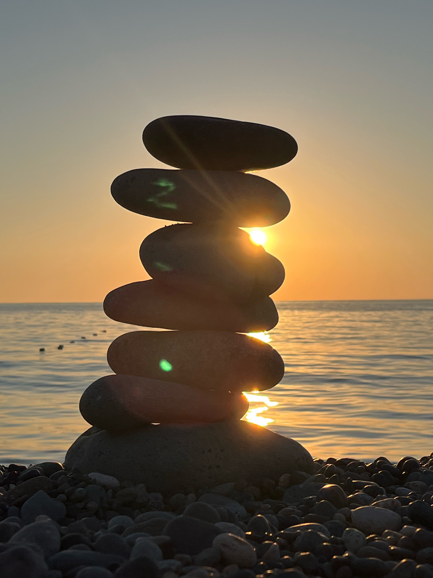 A stack of flat, dark stones balances precariously on a pebble beach, silhouetted against a vibrant sunset over the Black Sea. The sun's rays pierce through gaps in the stones, creating a bright glare and casting reflections on the water's surface. Some of the stones have faint green markings. In the distance, a line of buoys marks a boundary in the calm sea. The scene is set in Kobuleti, Georgia, a coastal city known for its pebble beaches and resorts. The time of day is dusk, indicated by the warm, golden hues of the sunset.