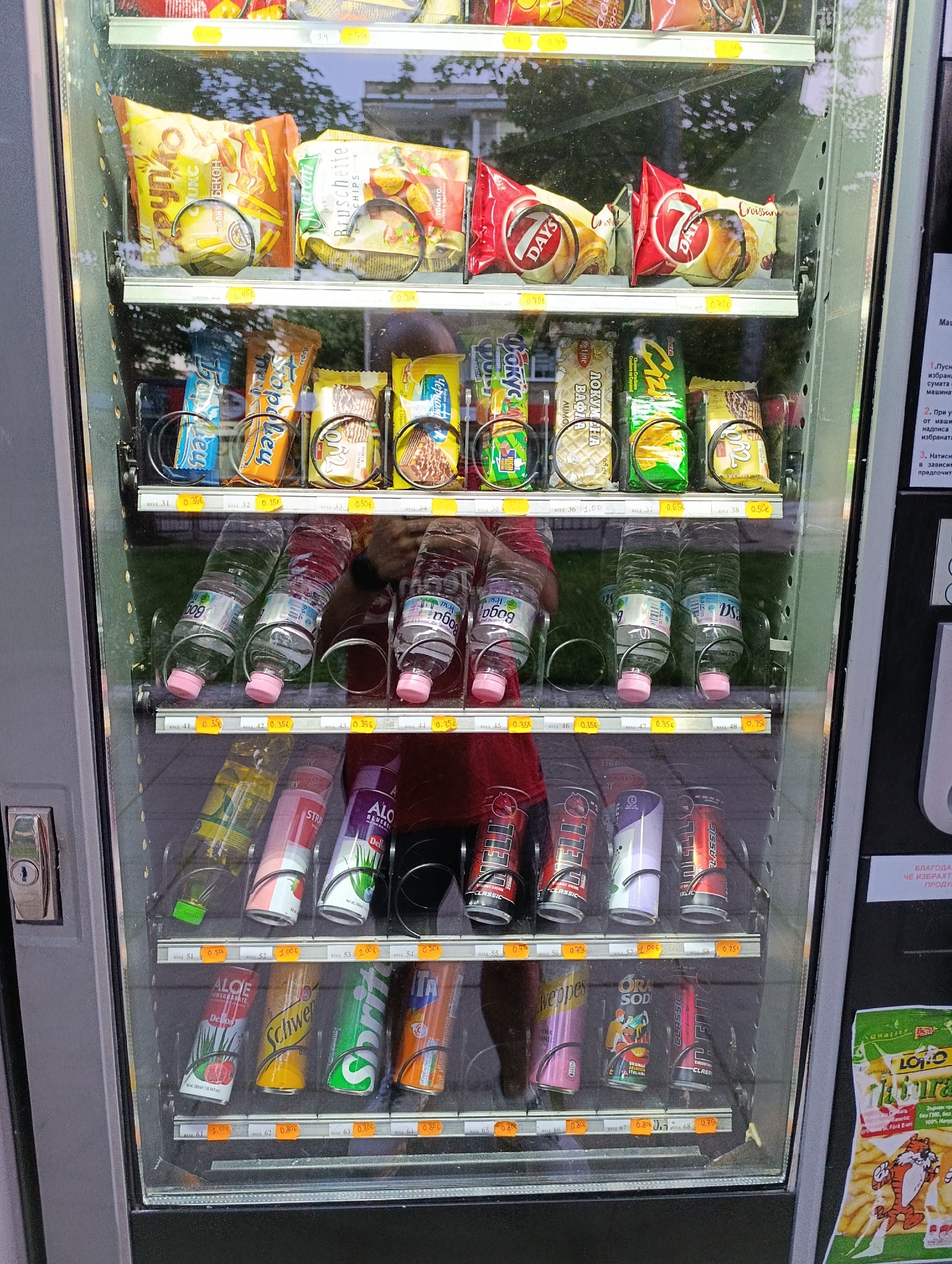 The image shows an exterior view of a vertical vending machine filled with various snacks and beverages. The machine has a glass front, displaying multiple rows of products. The top rows contain bagged snacks like chips and crackers. Below these, there are rows of bottled drinks and canned beverages. Some products are partially obscured by the internal mechanisms of the machine. The machine is located in an outdoor setting, with trees and foliage visible through the glass. A portion of a sign with text in an unfamiliar script is visible on the right side of the vending machine. The lighting suggests it is daytime.