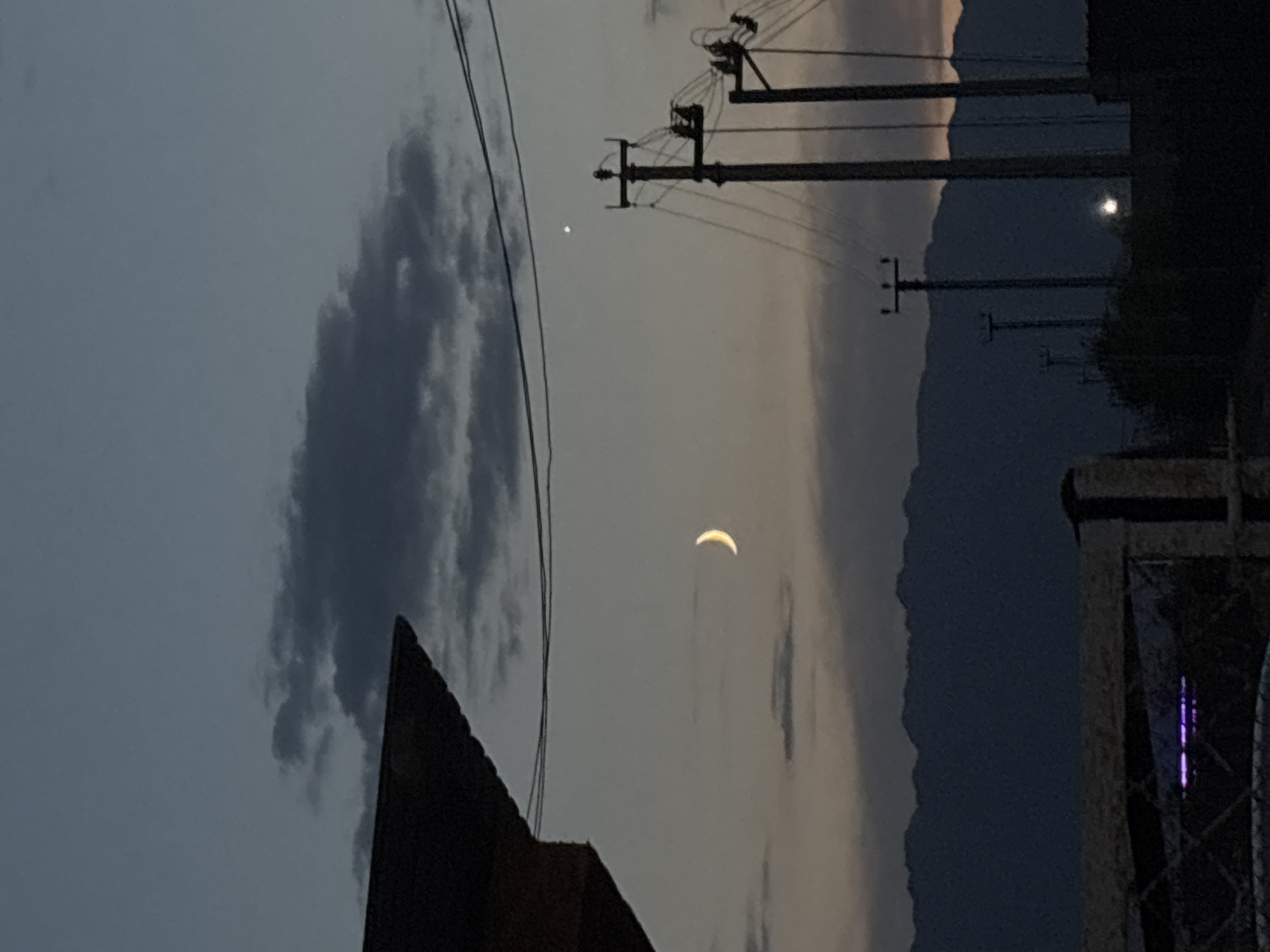 The image captures a twilight scene in Ақши, Kazakhstan, dominated by a dusky sky with scattered dark clouds. A sliver of the moon is visible in the upper left quadrant, accompanied by a bright, distant star or planet to its right. In the mid-ground, several utility poles with wires stretch across the frame, silhouetted against the fading light. Behind these structures, a range of dark, rugged mountains forms the horizon. In the foreground, the edge of a dark roof juts in from the left, and a fence runs horizontally across the bottom, with some indistinct objects and structures visible beyond it, including what appears to be a light emitting purple light. The overall atmosphere is subdued, characteristic of the transition from day to night.