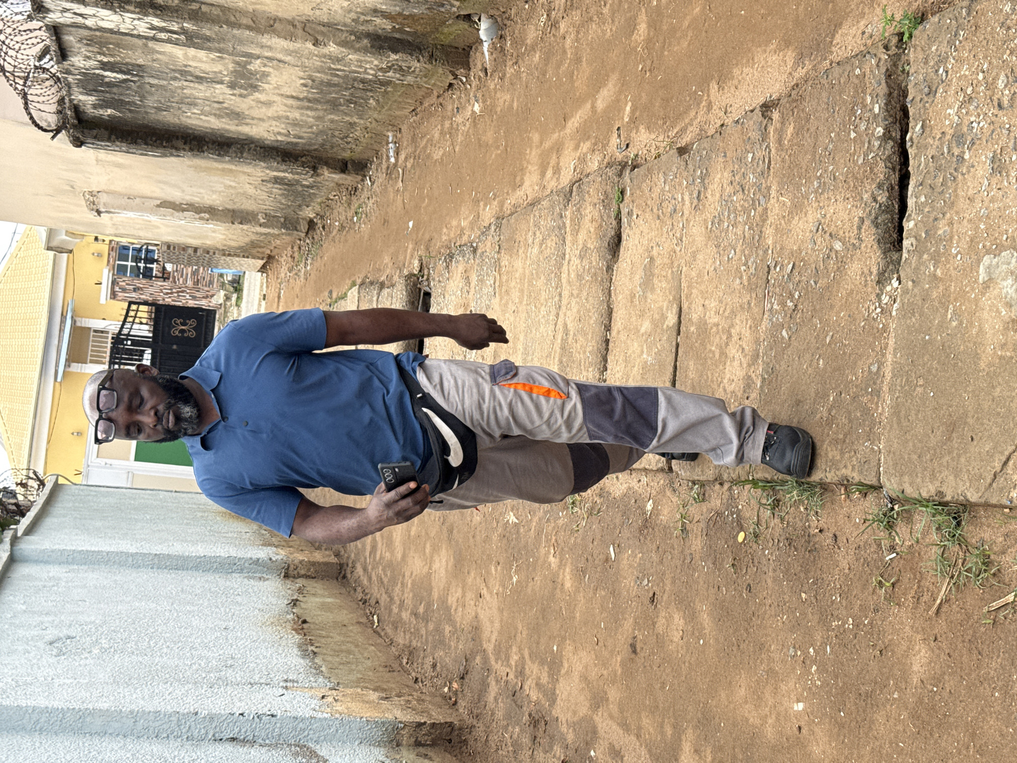 A medium, outdoor shot captures a man in a blue short-sleeved polo shirt, grey work pants with orange accents, and black shoes, standing on a narrow, dilapidated walkway. He is holding a smartphone in his left hand. The walkway is flanked by tall, weathered concrete walls, with vegetation growing in cracks and along the edges. The surface of the walkway is uneven and appears to be made of large, cracked concrete slabs. In the background, a yellow building with a black gate is visible. Barbed wire is visible atop a wall on the left. The presence of modern buildings, manicured lawns, and infrastructure indicate the setting is likely in a residential or commercial area of Abuja, Nigeria.
