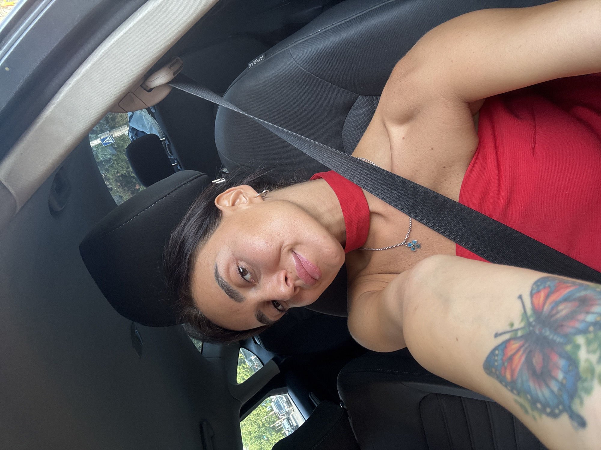 In a self-taken photograph, a woman is seated in the driver's side of a car. She is wearing a strapless red top and a red choker necklace, along with a silver necklace featuring a blue and silver cross pendant. Her left arm, visible in the foreground, is adorned with a colorful butterfly tattoo. The car's interior is dimly lit, with the black seatbelt prominently crossing her chest. Through the car windows, a street scene is visible, suggesting the location is Tbilisi, Georgia, indicated by the presence of trees, buildings, and other vehicles.