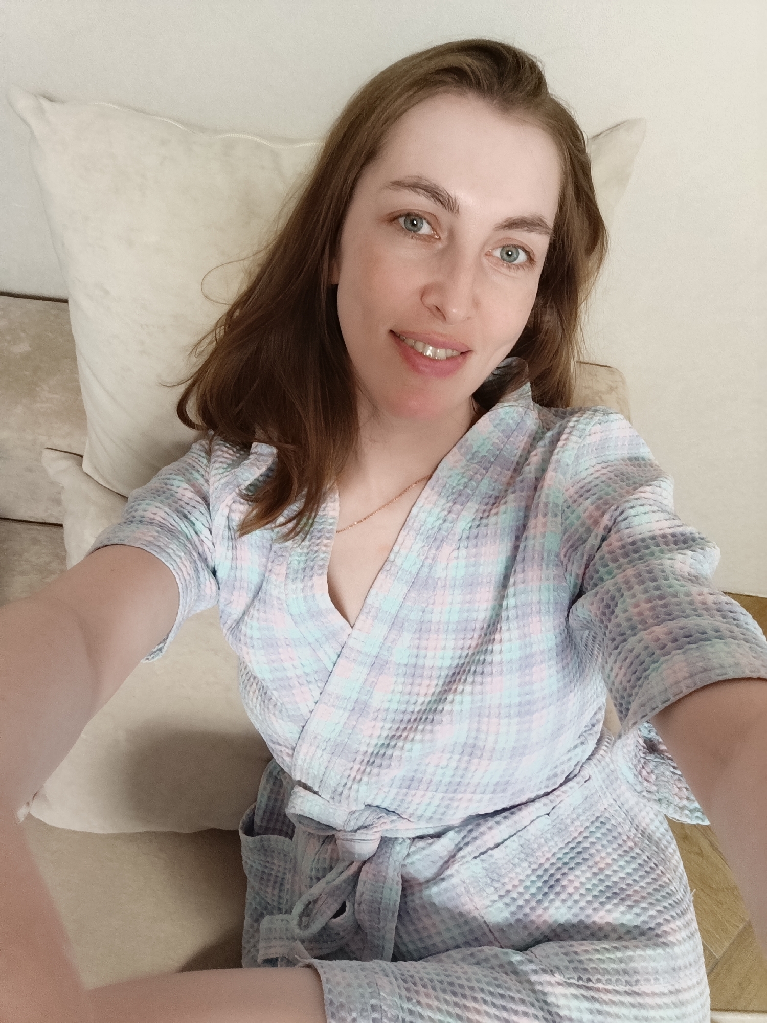 A woman is lying on a beige sofa with a beige pillow behind her. She is wearing a light blue, pink, and gray plaid bathrobe. She has light brown hair, blue eyes, and is smiling. She is wearing a gold necklace. The location is Chaikovsky, Russia.