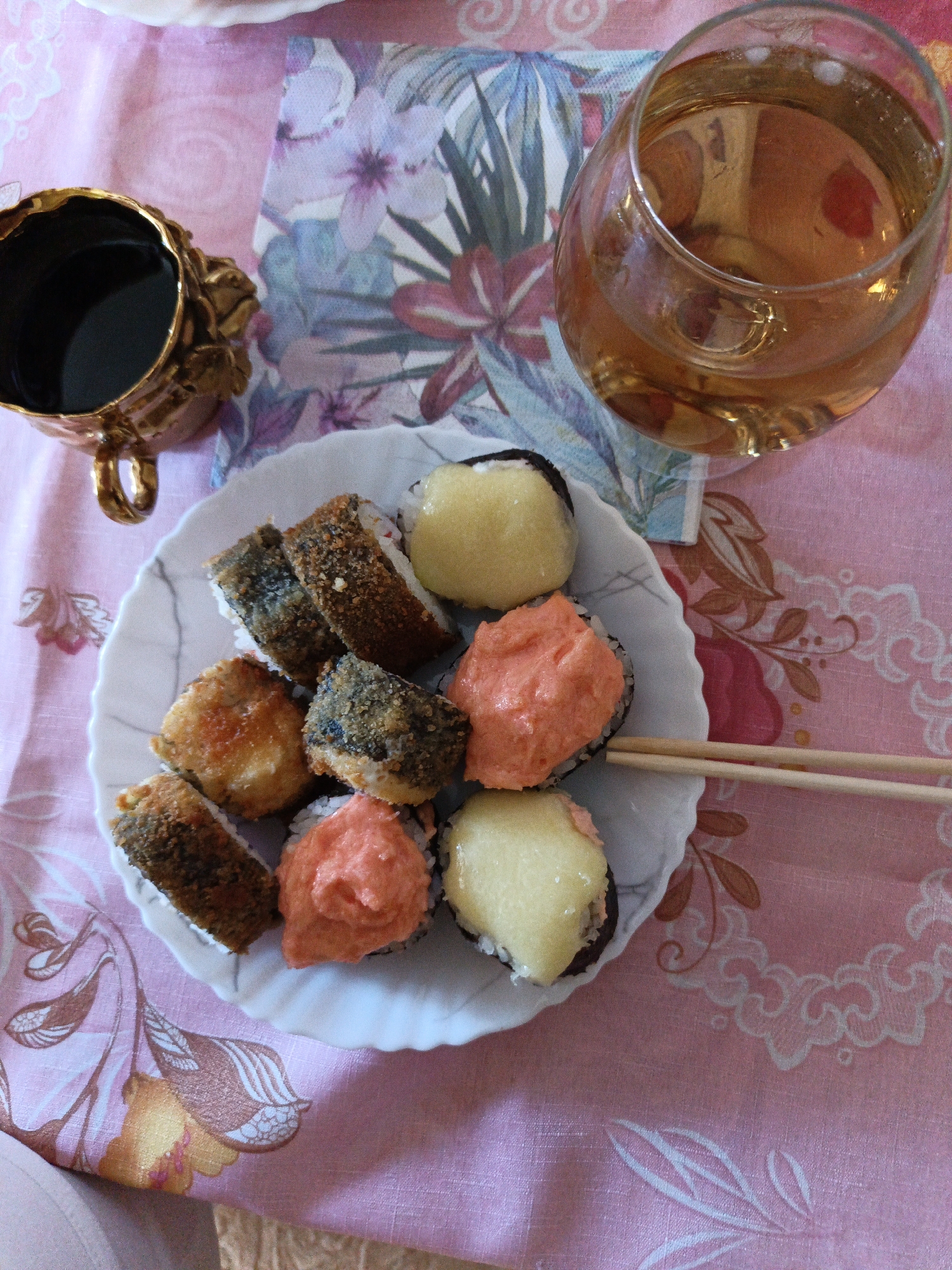 An overhead, eye-level shot shows a plate of sushi on a pink floral tablecloth, next to a golden ornate coffee cup and a glass of amber-colored liquid. The sushi is arranged in a circle on a white, scalloped-edge plate. There are at least eight pieces of sushi, some topped with what appears to be pink roe and others with a yellow creamy topping. Two wooden chopsticks rest on the plate, pointing towards the sushi. The coffee cup is made of a dark metal, possibly brass, and has an intricate, embossed floral design. The glass is transparent and slightly tilted, revealing the warm amber hue of its contents. A floral patterned napkin is visible beneath the glass and part of the plate. The tablecloth has a faded pink background with a repeating pattern of stylized pink and brown floral elements. The image does not contain any information that would allow for the determination of its location in the city of Novokuznetsk, Russia. There are no people visible in the image, and no actions or interactions are depicted beyond the placement of the sushi and chopsticks.