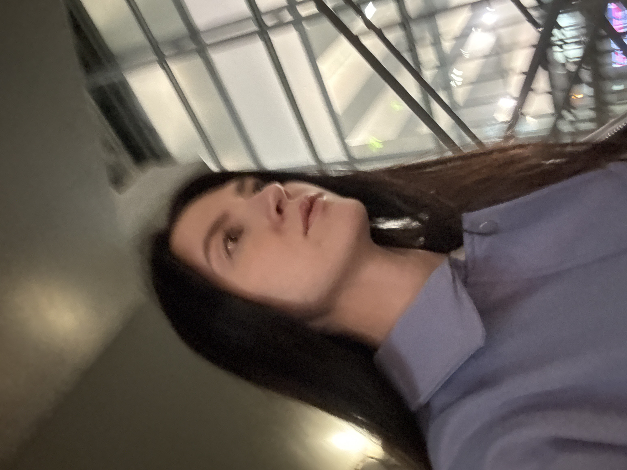 A woman with dark hair is looking up and to the left, seemingly inside a building with a glass ceiling, based on the beams and patterns visible above. She is wearing a collared shirt. The background suggests the interior of a modern structure. While it is unclear if it is Moscow, Russia, no conclusive evidence exists to confirm that location.