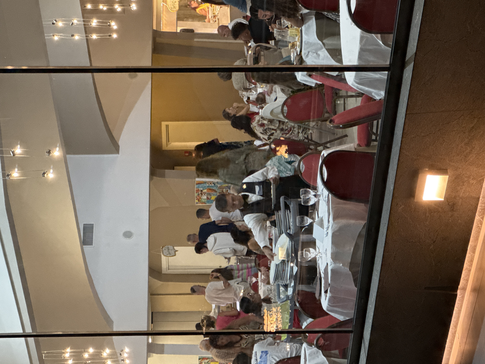 The image shows a busy restaurant scene, possibly in Capaci, Italy, viewed through a large window. Several tables are set with white tablecloths and red chairs, some occupied by diners. A waiter in a black vest and bow tie is seen carrying a plate of food, interacting with a stack of clean plates. In the background, more patrons are visible, some standing and conversing, while others are seated at tables. The restaurant appears to have a high ceiling with decorative light fixtures. The lighting is warm and ambient, suggesting an evening setting.