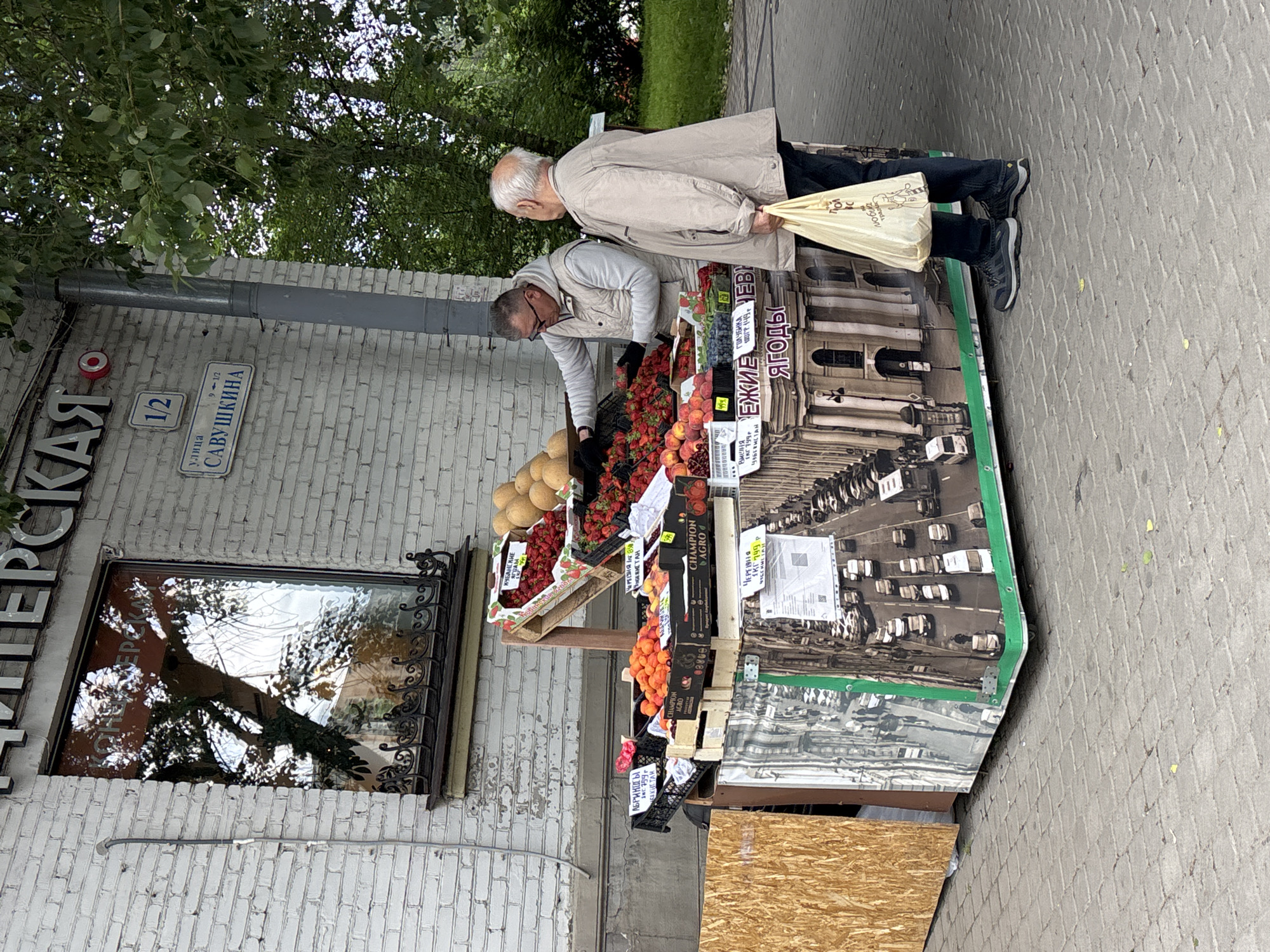 A street scene in Saint Petersburg, Russia, shows an outdoor fruit stand set up against a white brick building. Two men are present, with one leaning over the stand, examining the fruit on display, and the other standing beside him, holding a tan-colored bag. The fruit stand contains various fruits such as melons, strawberries, peaches, apricots, and blueberries, with signs in Russian indicating the type and origin of the produce. The street address, "улица Савушкина, 1/2" (Savushkina Street, 1/2), is visible on a sign attached to the building. A window with a decorative wrought-iron frame is also present. The street surface is made of gray paving stones. The stand has a photograph of a building on its side.