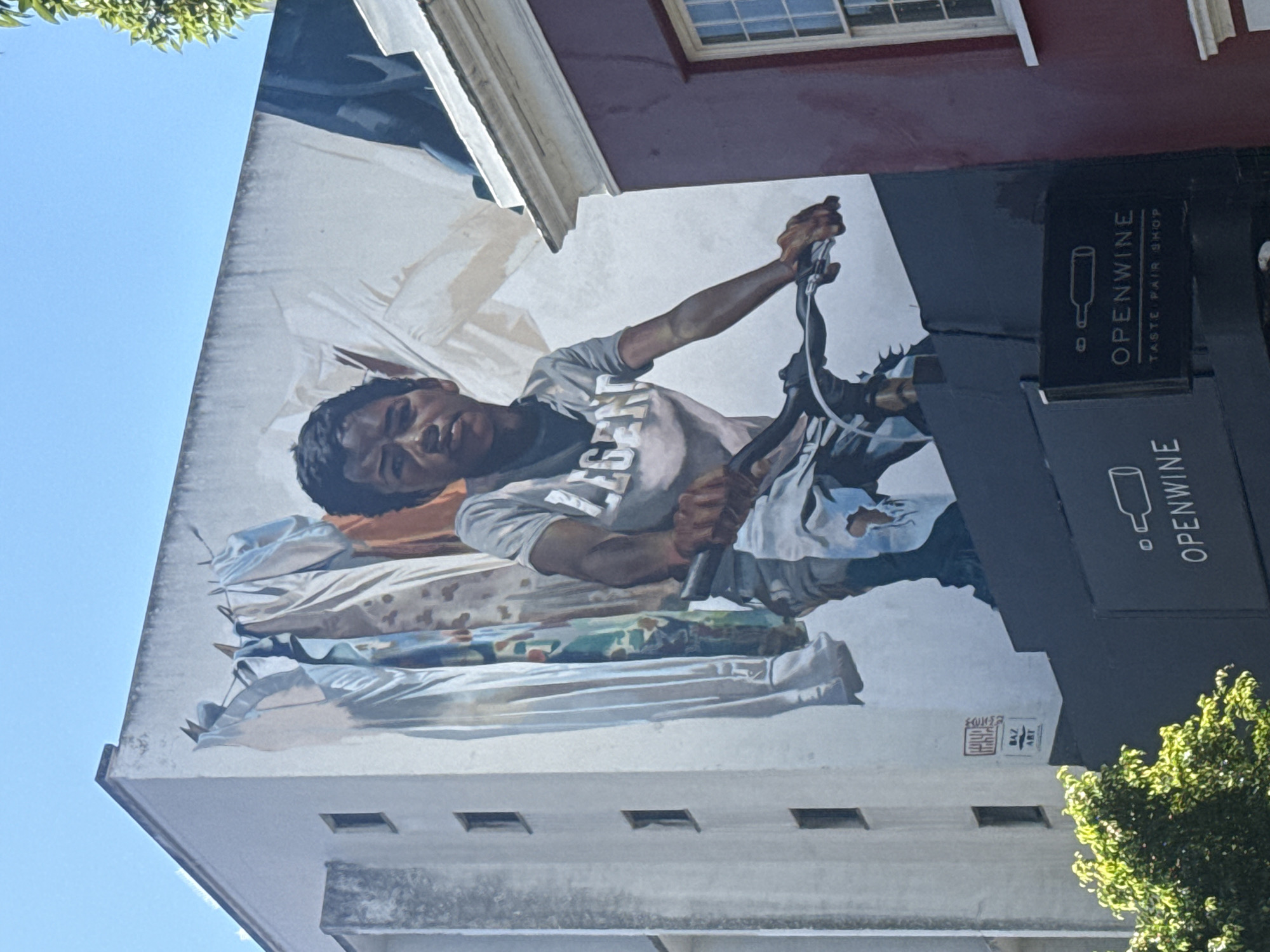 The image is a mural painted on the side of a building. The mural depicts a person with dark hair, wearing a grey t-shirt with white lettering, and holding a black object. The person is positioned above a stack of items covered in draped fabric, with colors of brown, blue, and white. The background is primarily white with hints of grey and light blue. A black sign for "OPENWINE TASTE PAIR SHOP" is visible to the right. A signature is on the bottom-right corner of the mural. The building is multi-story and a corner of a window frame is at the top of the image. The sky is blue, and portions of green foliage are visible in the upper-left and bottom-right corners. This location is in Cape Town, South Africa.