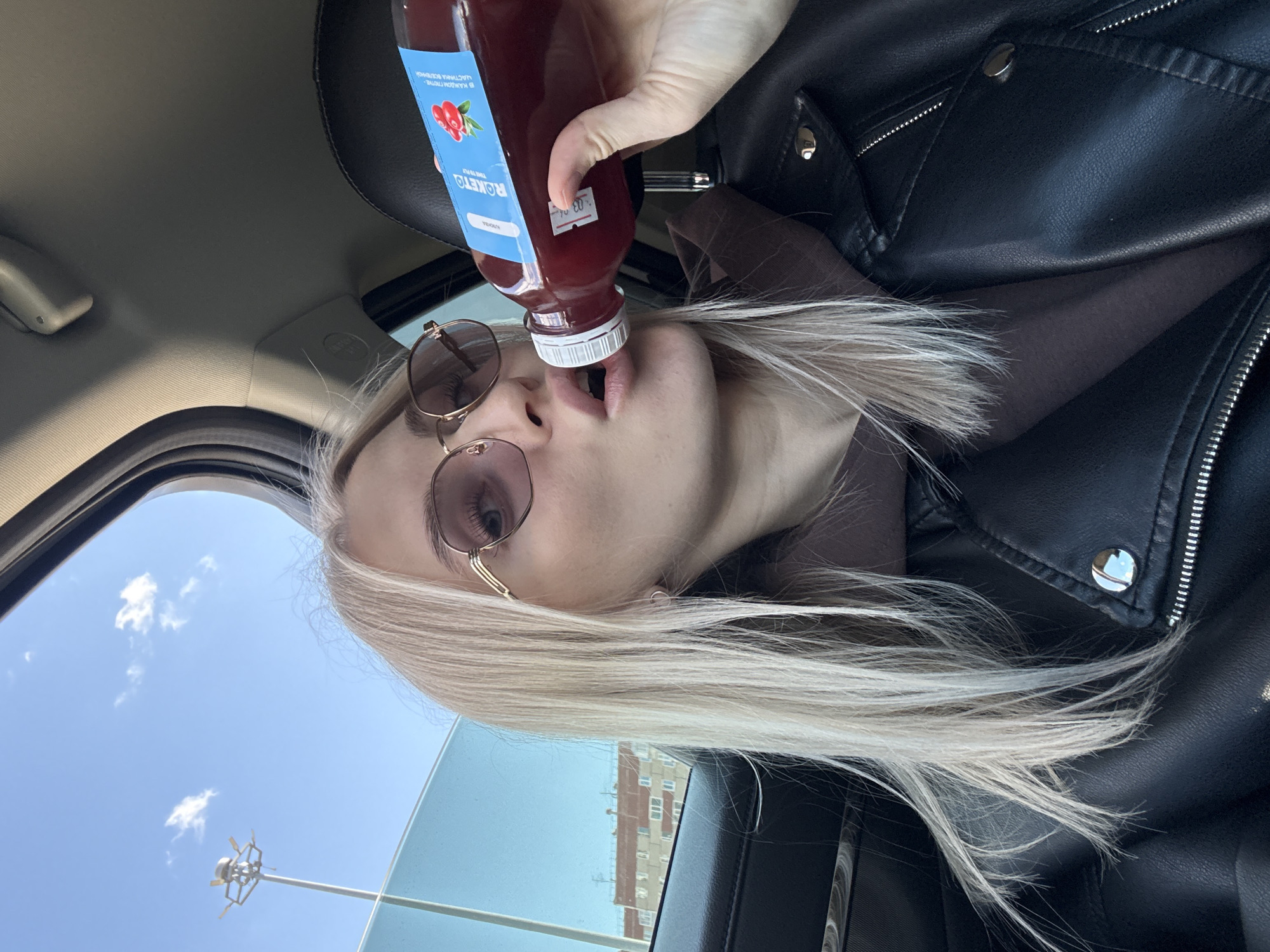 A woman with platinum blonde hair, wearing rose-tinted sunglasses and a black leather jacket, is drinking from a bottle of lingonberry juice while sitting in a car. The label on the bottle reads "ROKETO", with the words "LINGONBERRY FLAVORED IN EVERY SIP" in Russian. Outside the car window, a light-blue sky is visible, along with a street lamp and the facade of an apartment building, suggesting a location in Izhevsk, Russia.
