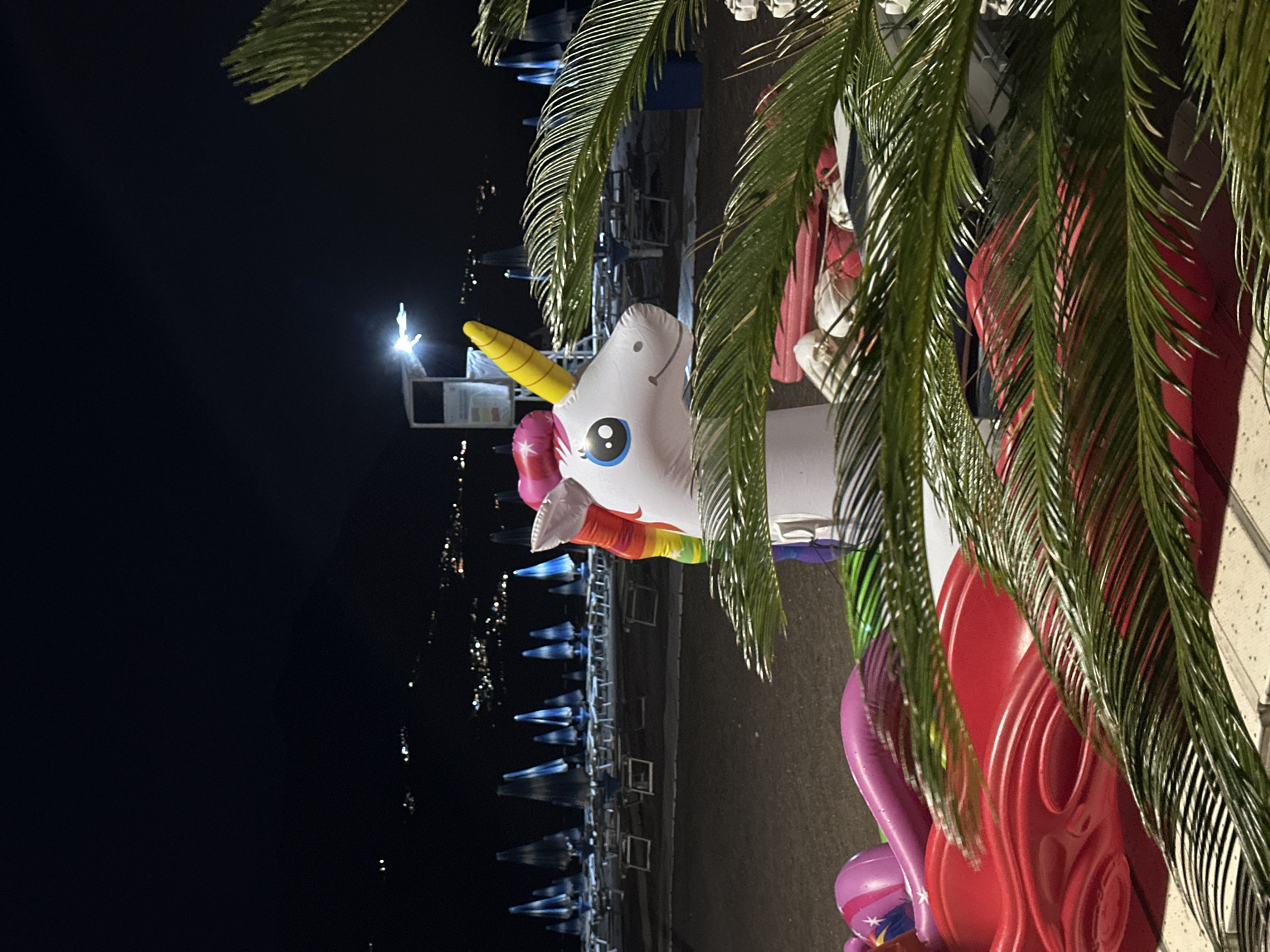 The image captures an inflatable unicorn positioned at what appears to be a nighttime event or attraction in Boreti, Montenegro. The unicorn is white with a yellow horn, blue eyes, and a rainbow-colored mane. It is partially obscured by lush green palm fronds, which frame the scene and add a tropical ambiance.
In the background, rows of empty beach chairs are set up, suggesting a beach or waterfront location. The chairs are covered and arranged neatly, hinting at the end of a day or preparation for the next. Beyond the chairs, a body of water is visible, reflecting numerous small lights, possibly from a distant town or boats. The overall impression is one of a serene, possibly festive, nighttime setting.
The time of day is clearly night, indicated by the darkness and the presence of artificial lights. The weather appears calm, with no strong winds suggested by the foliage. The scene evokes a sense of calm and perhaps anticipation for an event or activity. There is a small white structure with a spotlight in the background, which could be a lifeguard station or a booth for information. No visible text is discernible in the image. The presence of the inflatable unicorn and the beach chairs suggests a recreational or entertainment-focused location, likely a resort or public beach area.