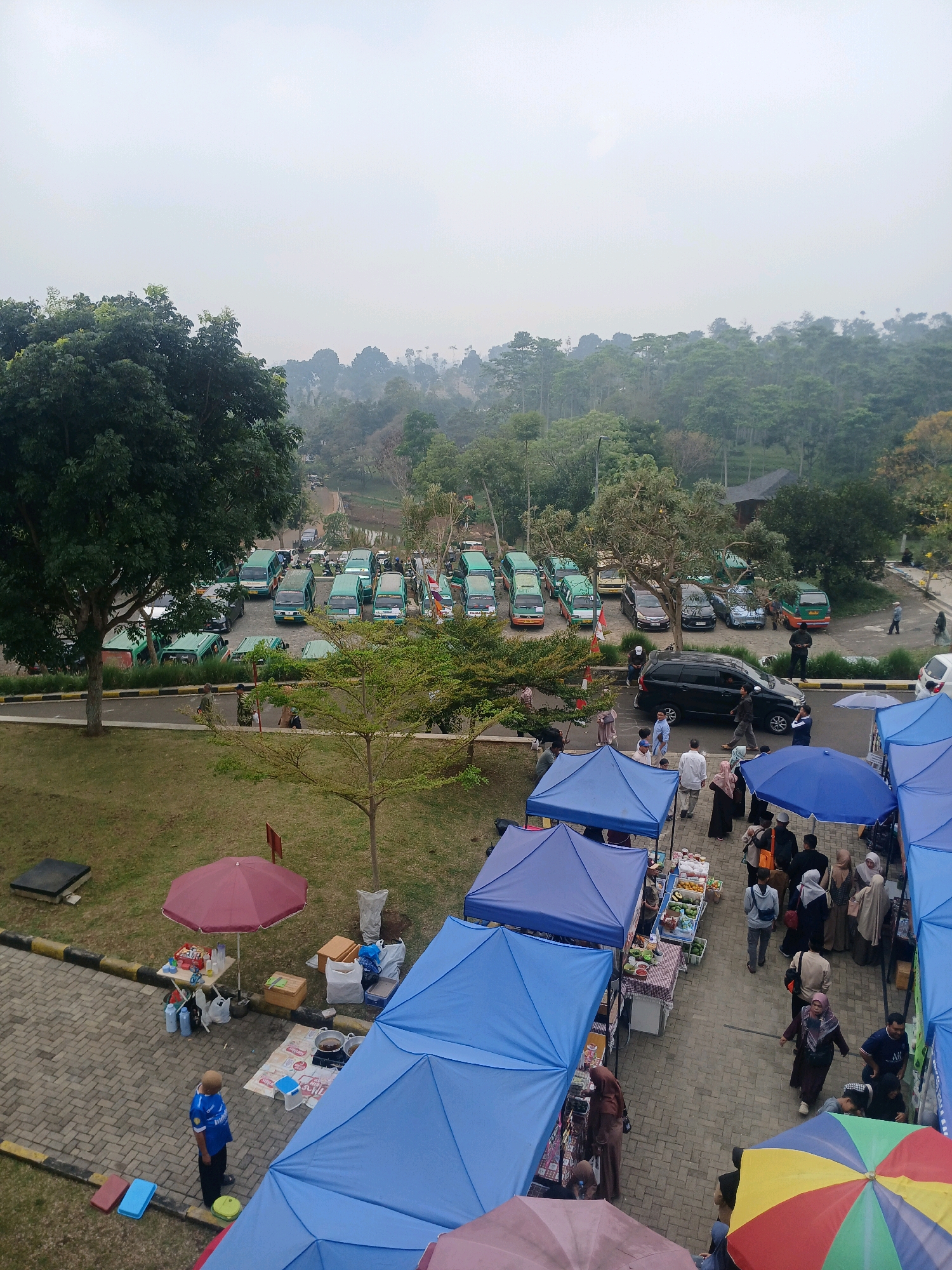 The image depicts an outdoor market scene likely located in Arjasari, Indonesia. In the background, numerous green vans are parked in a lot, suggesting a transportation hub or a large gathering. A dense forest with a hazy sky forms the backdrop, indicating a potentially rural or semi-rural environment.
In the foreground, a marketplace is set up with several blue and purple tents. Vendors are visible behind tables displaying various goods, including what appear to be fruits and food items. Shoppers, mostly women dressed in headscarves, are browsing the stalls and interacting with vendors. Some individuals are walking, while others stand and observe the market. A few red and yellow umbrellas are also present, providing shade.
One stall features a cooking setup with a pot on a stove. A man in a blue shirt stands near a maroon umbrella, looking towards the market. The ground is paved with brick-like material, and grassy areas are interspersed with the market stalls. The overall atmosphere suggests a busy, community-oriented event.