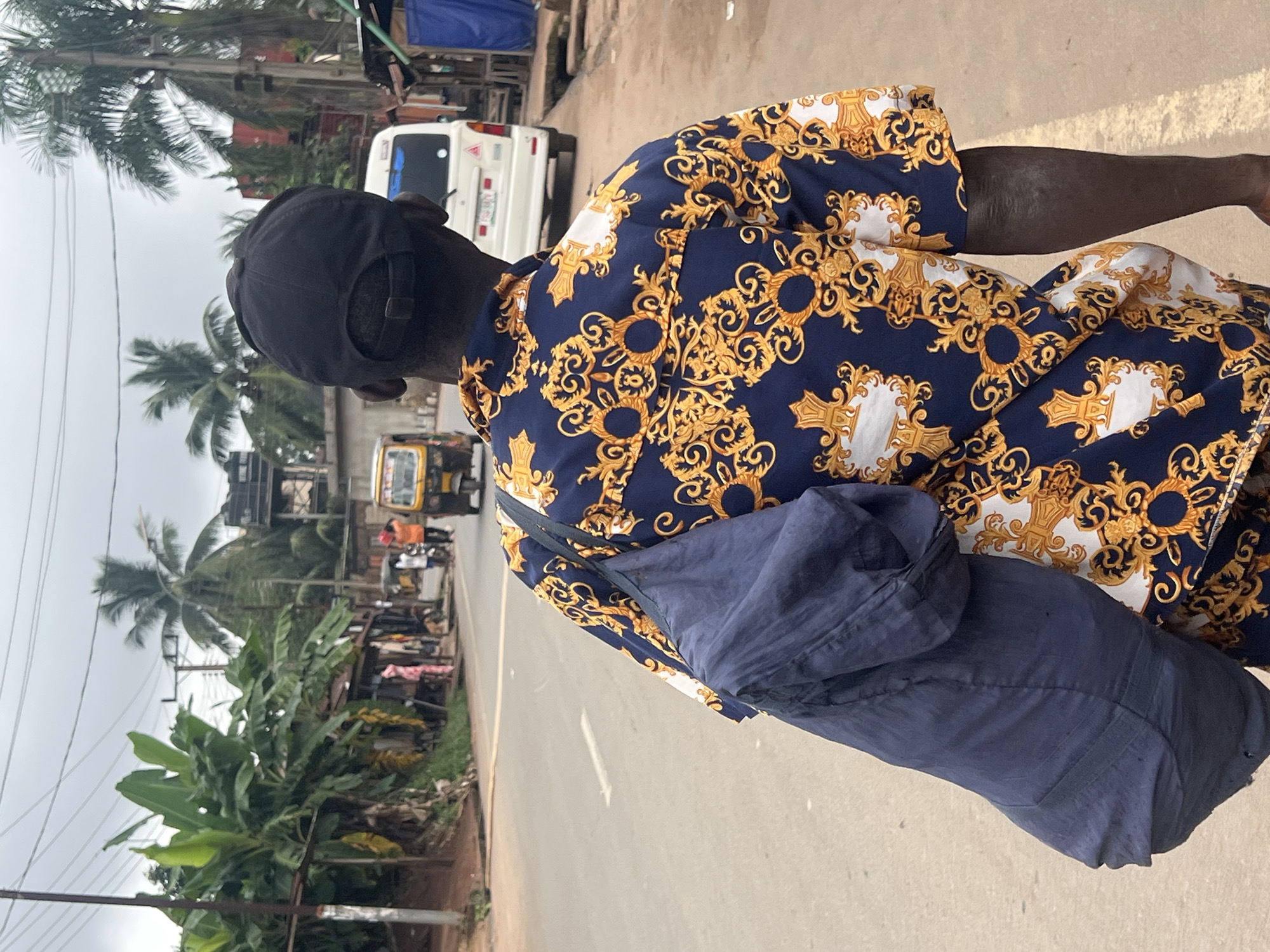 A person is walking away from the camera on a paved road in Nkpor, Nigeria. They are wearing a dark blue and gold patterned short-sleeved shirt and a black baseball cap. A dark blue bag is slung over their left shoulder. The road is lined with buildings and vegetation, including banana trees and palm trees. Several vehicles are visible on the road, including a yellow auto-rickshaw and a white van. The sky is overcast.