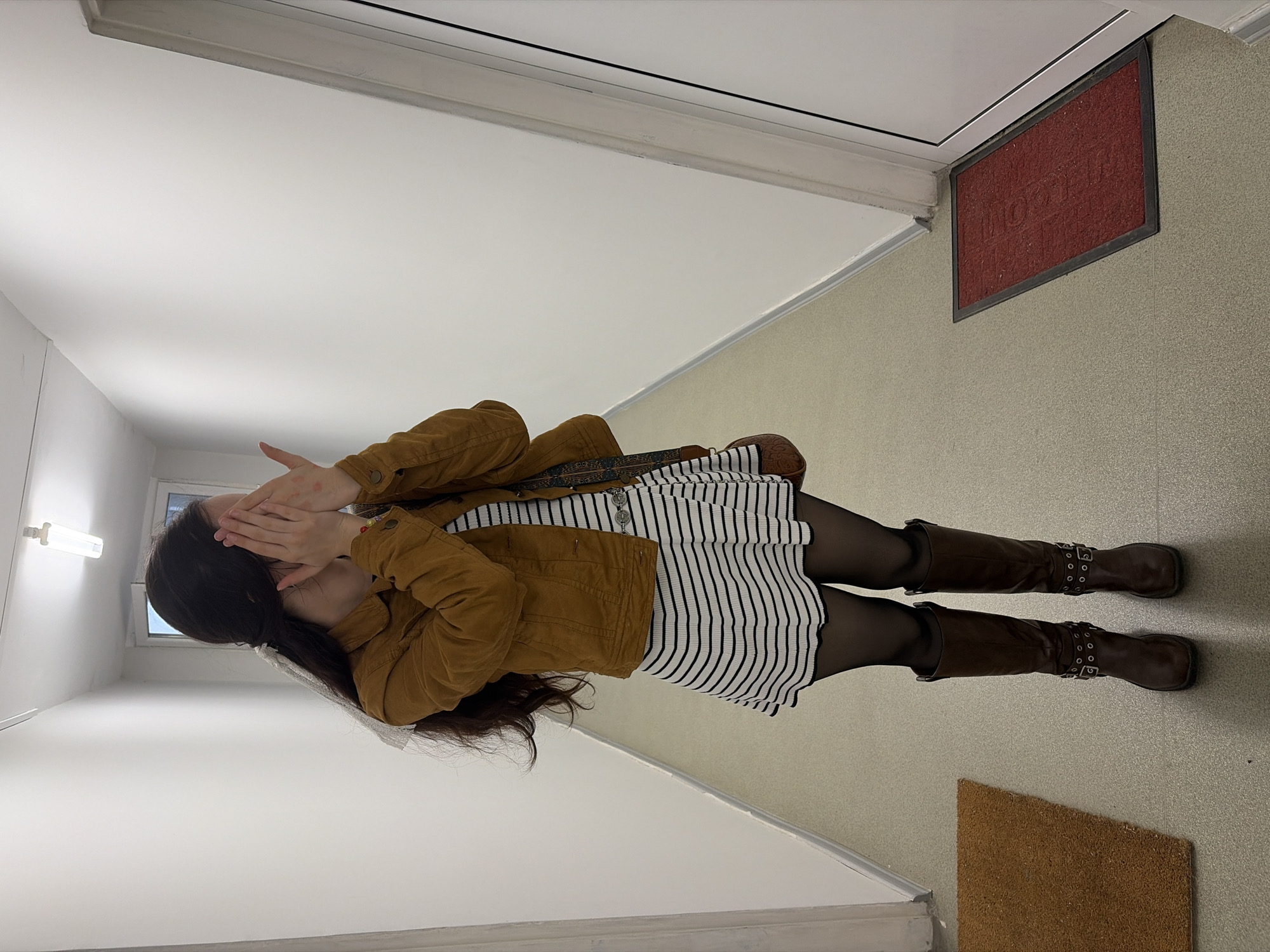 The media file features a young woman standing in an indoor hallway or corridor, appearing to be an apartment entrance. The image, originally shot in portrait, is oriented horizontally. She is covering her face with both hands, a gesture that suggests shyness, embarrassment, or a playful 'peeking'; red marks are visible on her right hand.
She is dressed in a brown jacket, which appears to be made of corduroy or a similar textured fabric. Beneath the jacket, she wears a white dress or skirt with black horizontal stripes. Her legs are covered in black opaque tights, and she is wearing tall, dark brown boots with multiple decorative buckles and straps. A patterned strap, likely from a handbag, is visible over her shoulder. Her long, dark hair is pulled back, fastened with a white ribbon or hair tie.
The setting is brightly lit, with white walls on one side and a lighter, possibly beige or light grey textured wall on the other. The floor is a light, neutral color. A simple fluorescent-style light fixture is mounted on the white wall, and a window in the background indicates it is daytime.
Notable objects include a red rectangular mat with the word "WELCOME" clearly embossed on it, which is unusually mounted on the textured wall near a white door frame. A smaller, brown textured mat is also visible on the floor. The overall scene suggests a clean, residential interior, consistent with a location like Sofia, Bulgaria.