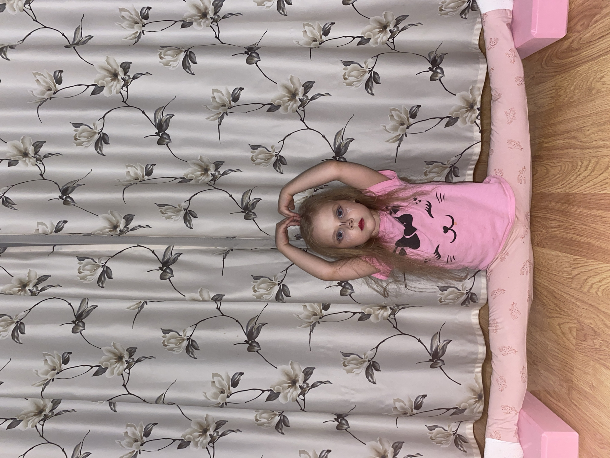 A young child is captured performing a full split on a patterned fabric surface in an indoor setting, likely in Votkinsk, Russia. The child, with blonde hair, wears a pink t-shirt with a black cat face design and pink leggings featuring unicorn patterns. She lies horizontally across the frame, with her legs fully extended to the left and right. Her arms are raised above her head, forming a heart shape with her hands. She looks directly at the camera with a neutral expression, wearing white glittery eye makeup and red lipstick.
The surface beneath the child is a light-colored fabric, possibly a curtain or bedspread, adorned with a repeating floral pattern of cream-colored flowers and dark leaves on branches. The fabric shows horizontal pleats or folds. To the right of the child, a wooden floor is visible, alongside a partially seen pink object, possibly furniture or a toy. No text is visible in the image.