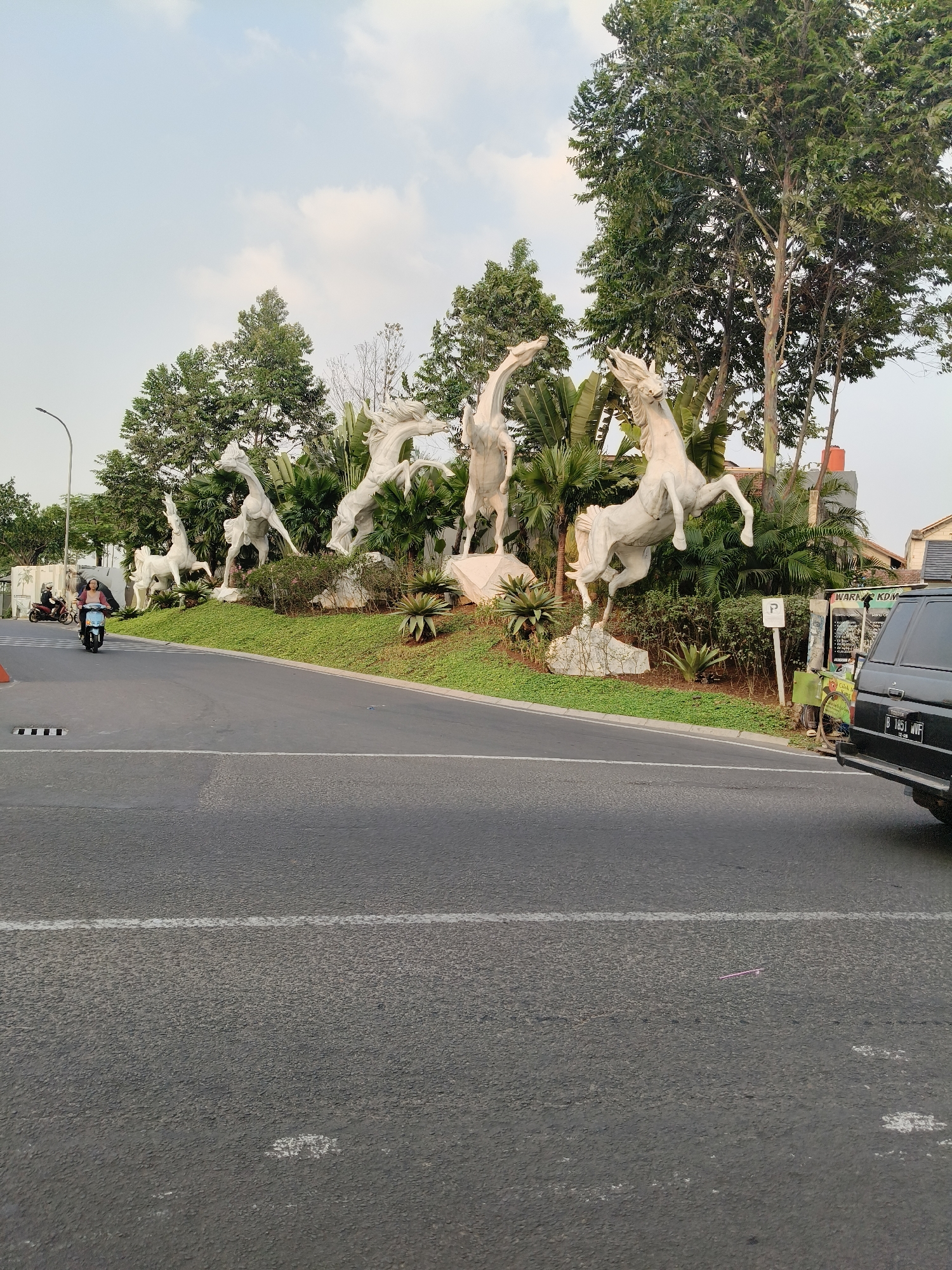 The image shows a roadside scene in South Tangerang, Indonesia, featuring a sculptural display of white horses against a backdrop of lush greenery and trees. In the foreground, a grey asphalt road is visible with white lane markings. A black SUV is parked on the right side of the road, with its rear visible. To the left, a person rides a light blue scooter, passing by the horse sculptures. Another person on a red motorcycle is also visible further to the left. A street lamp stands near the left edge of the image. The horse sculptures are dynamically posed, appearing to be in motion, and are positioned on a landscaped mound covered with grass and various plants. In the background, there are buildings and more trees, under a cloudy sky.