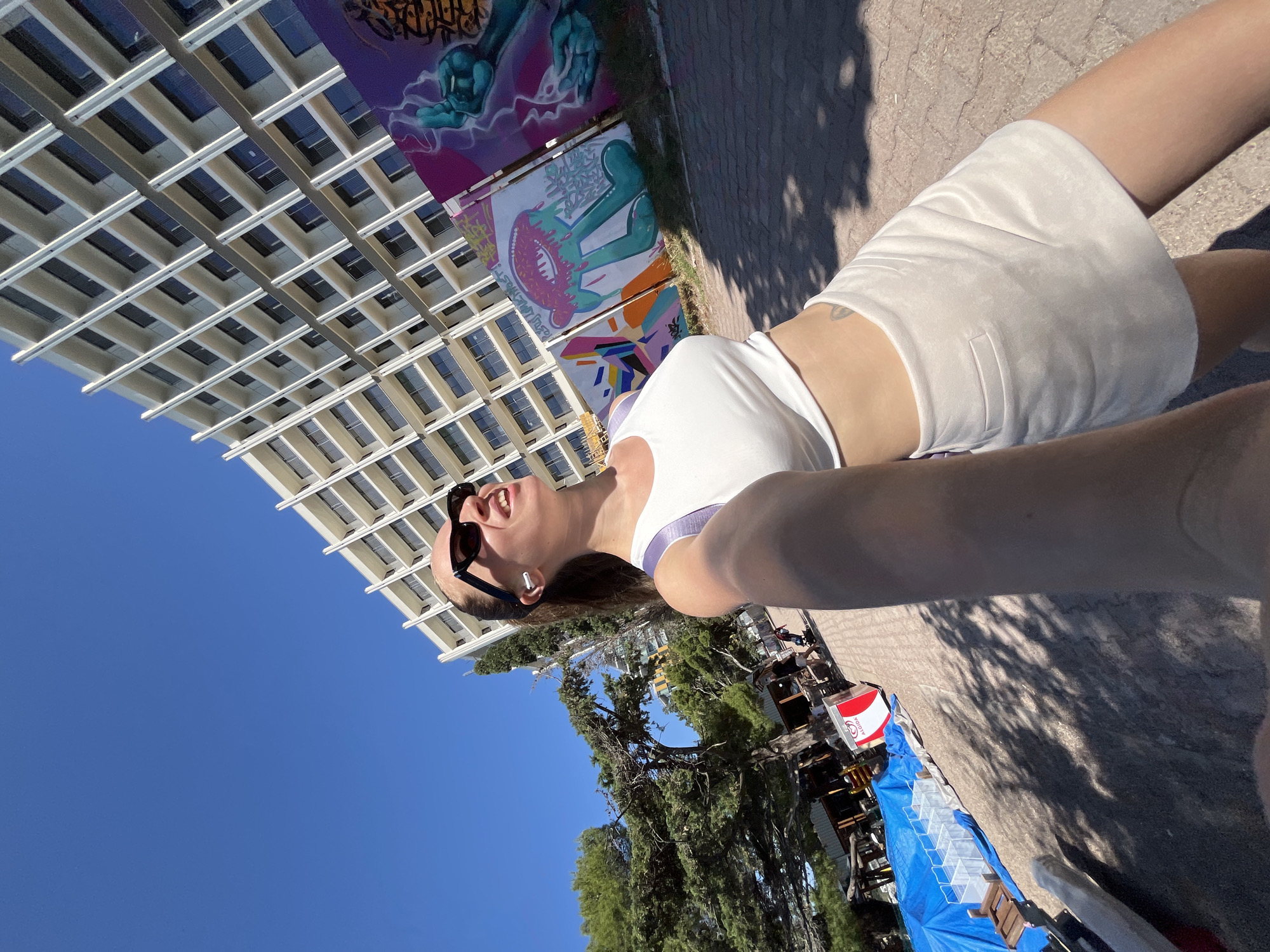 A selfie-style photograph features a woman against a backdrop of Rafailovići, Montenegro. The perspective is unusual, with the camera pointed upwards. The woman, wearing sunglasses, a white tank top, and a white skirt, is partially visible. The sky above is clear blue. Buildings, some adorned with murals, are visible. Additionally, trees, a street, and objects under a blue tarp are in view.