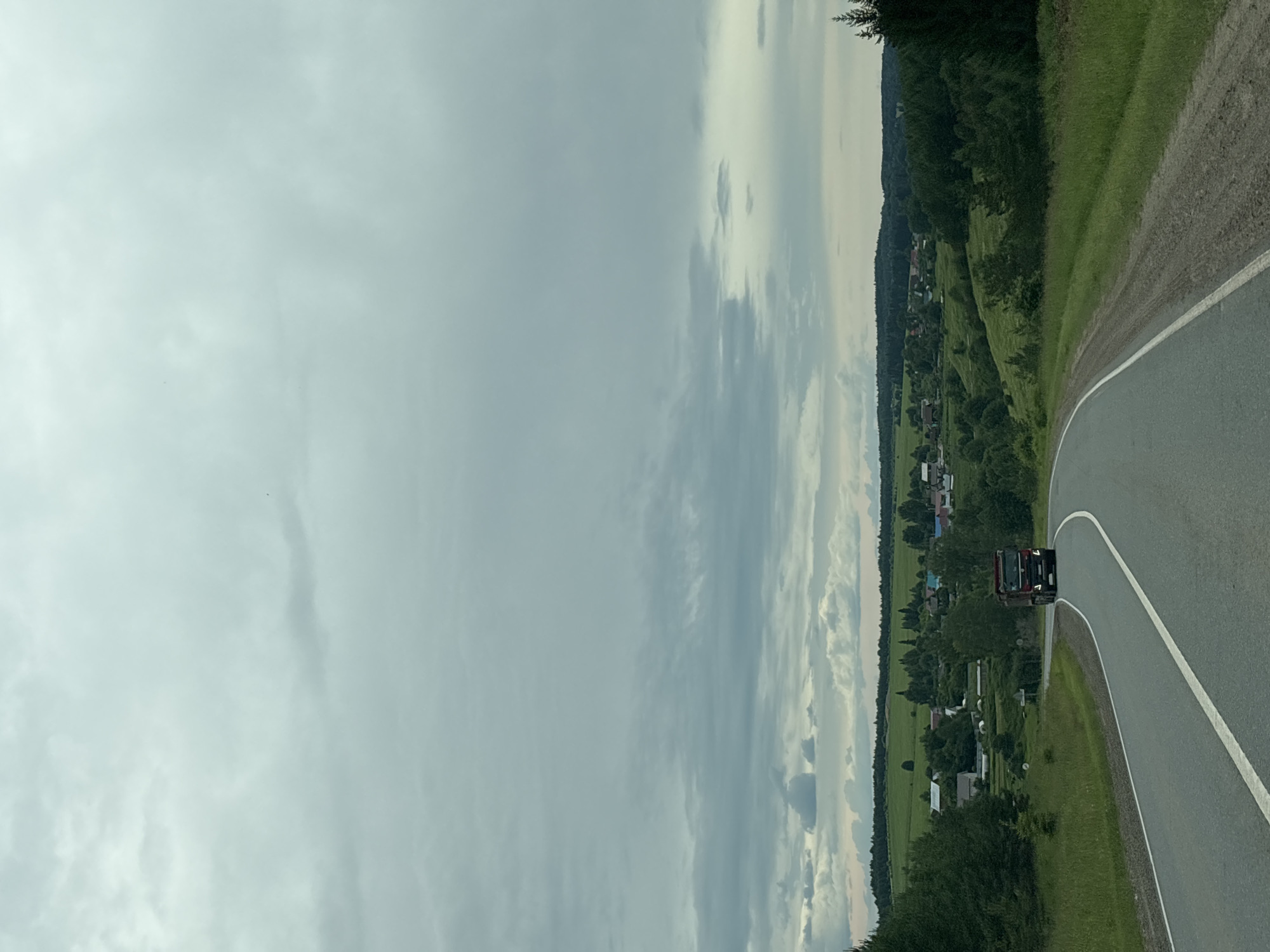 The image depicts a rural scene in Забегалово, Russia. A red truck is driving on a paved road that curves to the right. The road is bordered by a grassy embankment on the left and a dense green forest on the right. Beyond the embankment, a village is nestled in a valley. Houses with red and blue roofs are visible amidst lush green fields and trees. The sky is overcast with scattered clouds, suggesting a partly cloudy or grey day. The time of day appears to be daylight, possibly late afternoon or early evening given the lighting. The overall mood is serene and peaceful, capturing a typical rural landscape. There is no visible text in the image.