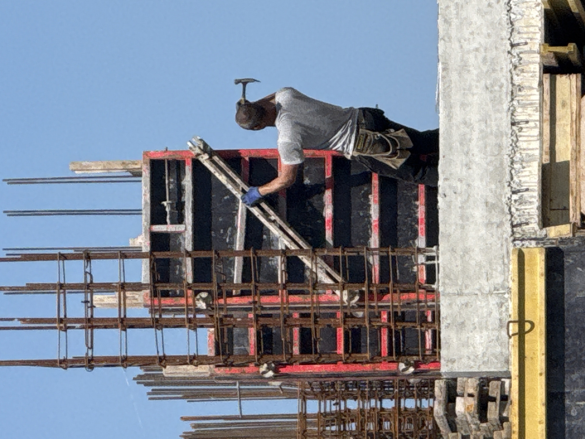 An outdoor daylight scene captures a construction worker engaged in building activity at a construction site in Varna, Bulgaria. The clear blue sky overhead indicates bright, sunny weather, likely midday or early afternoon.
A male construction worker is the central subject, dressed in a light grey short-sleeved t-shirt, dark trousers, and a tool belt around his waist. He wears a cap or head covering, and a blue glove is visible on his right hand. A hammer is tucked into the back of his collar, handle facing upwards. He is leaning forward over a section of concrete formwork, reaching down with his gloved hand, actively working on the structure.
The scene is dominated by construction materials. Extensive grids of rebar (reinforcing bars), some with visible rust, are prominent in the foreground and mid-ground, indicating a reinforced concrete structure. Sections of black and red framed concrete formwork are assembled around the rebar, ready for concrete pouring. To the right, a freshly poured, light grey concrete column or wall section is visible, next to a tall yellow support beam featuring a small metal handle. White plastic spacers are also discernible among the rebar.
The image provides a snapshot of active construction, showcasing the intricate framework of rebar and formwork being prepared or adjusted by the worker. No discernible text is visible in the image.