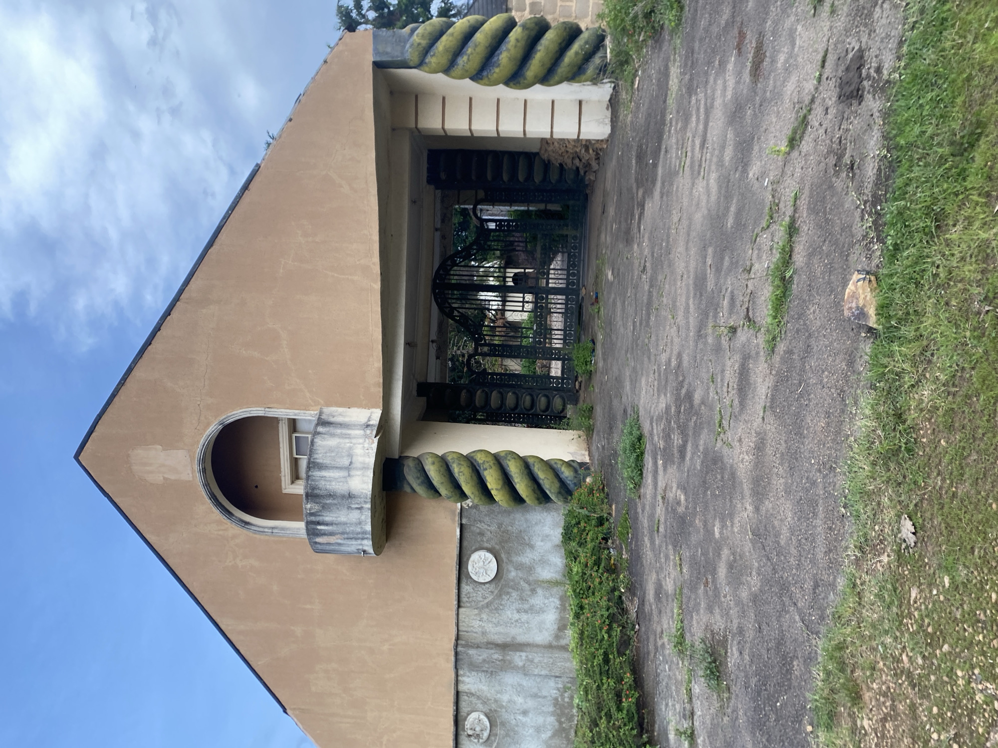 The image depicts the entrance to an abandoned property in Barikin Jirga Summa, Nigeria. The facade of the building is a tan color with a prominent arched balcony. Flanking the entrance are large, twisted columns with a green and yellow spiral design. A black wrought-iron gate stands open, revealing a glimpse of overgrown greenery beyond. The driveway is cracked asphalt, with patches of grass growing through. The overall scene suggests neglect and disuse, with a somber atmosphere under a cloudy sky. There are no people or animals visible in the image.